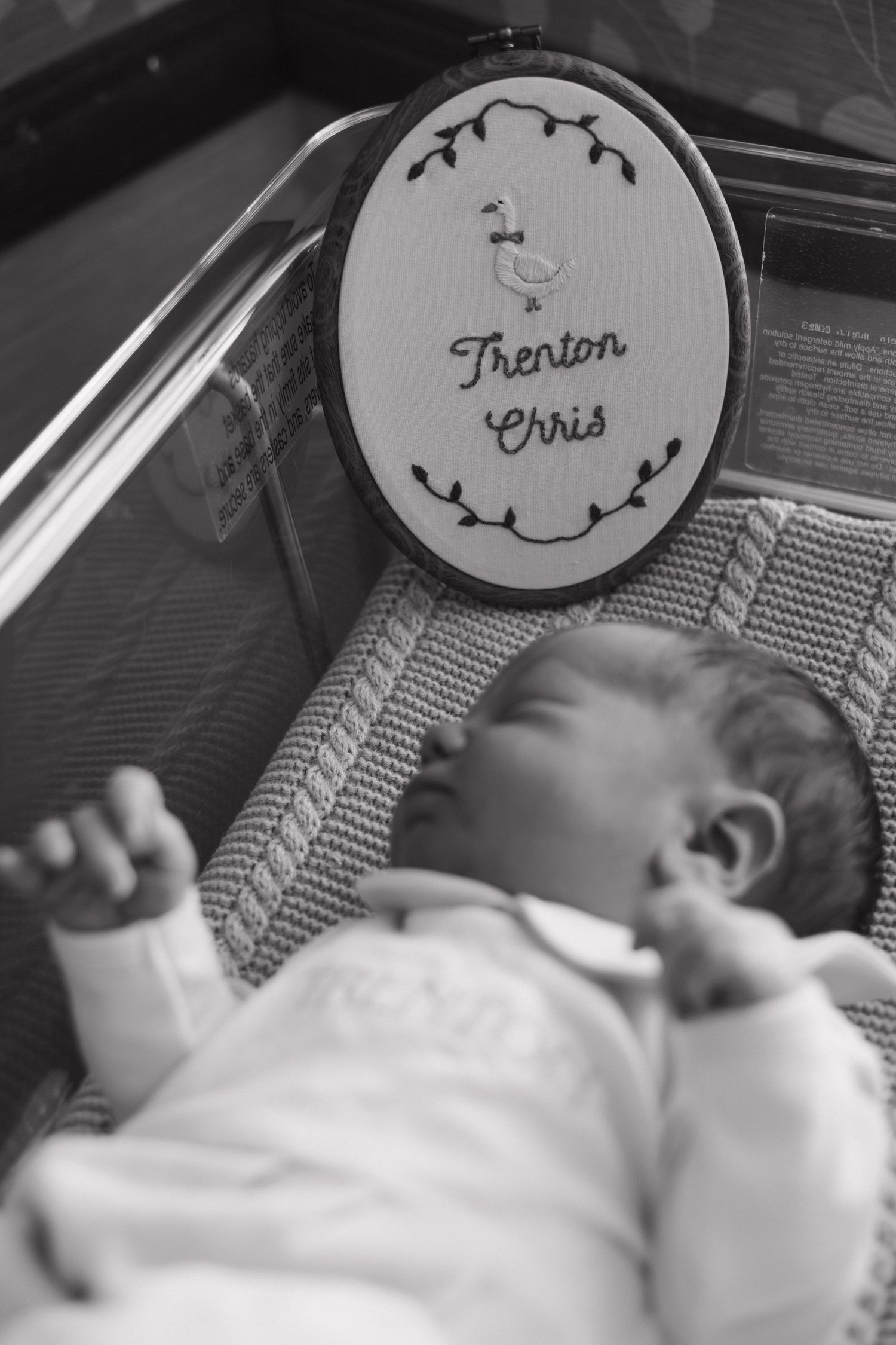 Black and white photo of newborn baby Trenton Chris lying in hospital bassinet with embroidered name sign.