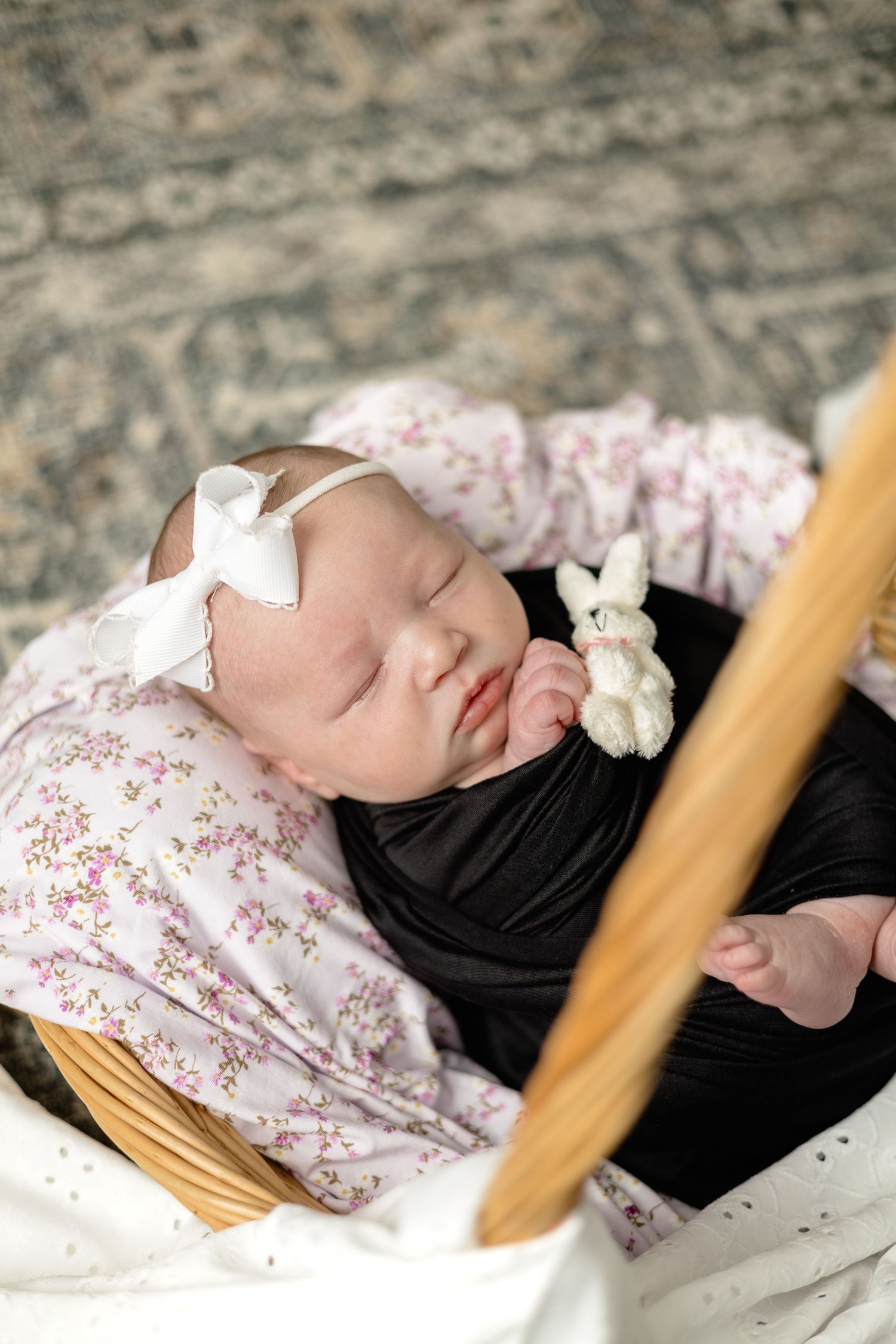 Sleeping newborn baby girl in a wicker basket, wrapped in black, wearing a white bow headband, holding a tiny stuffed bunny.