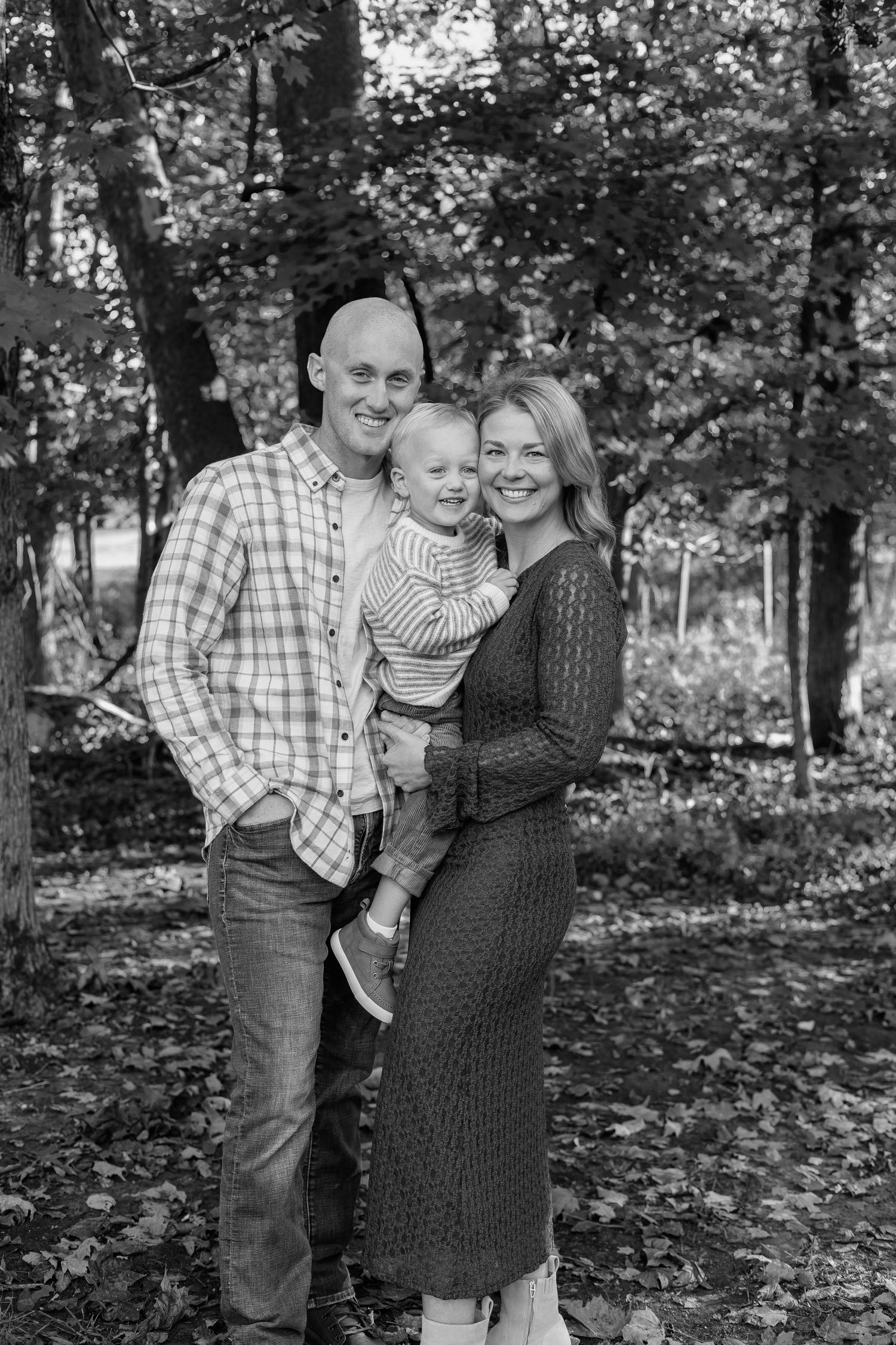 Black and white family portrait of parents holding toddler in autumn forest setting.