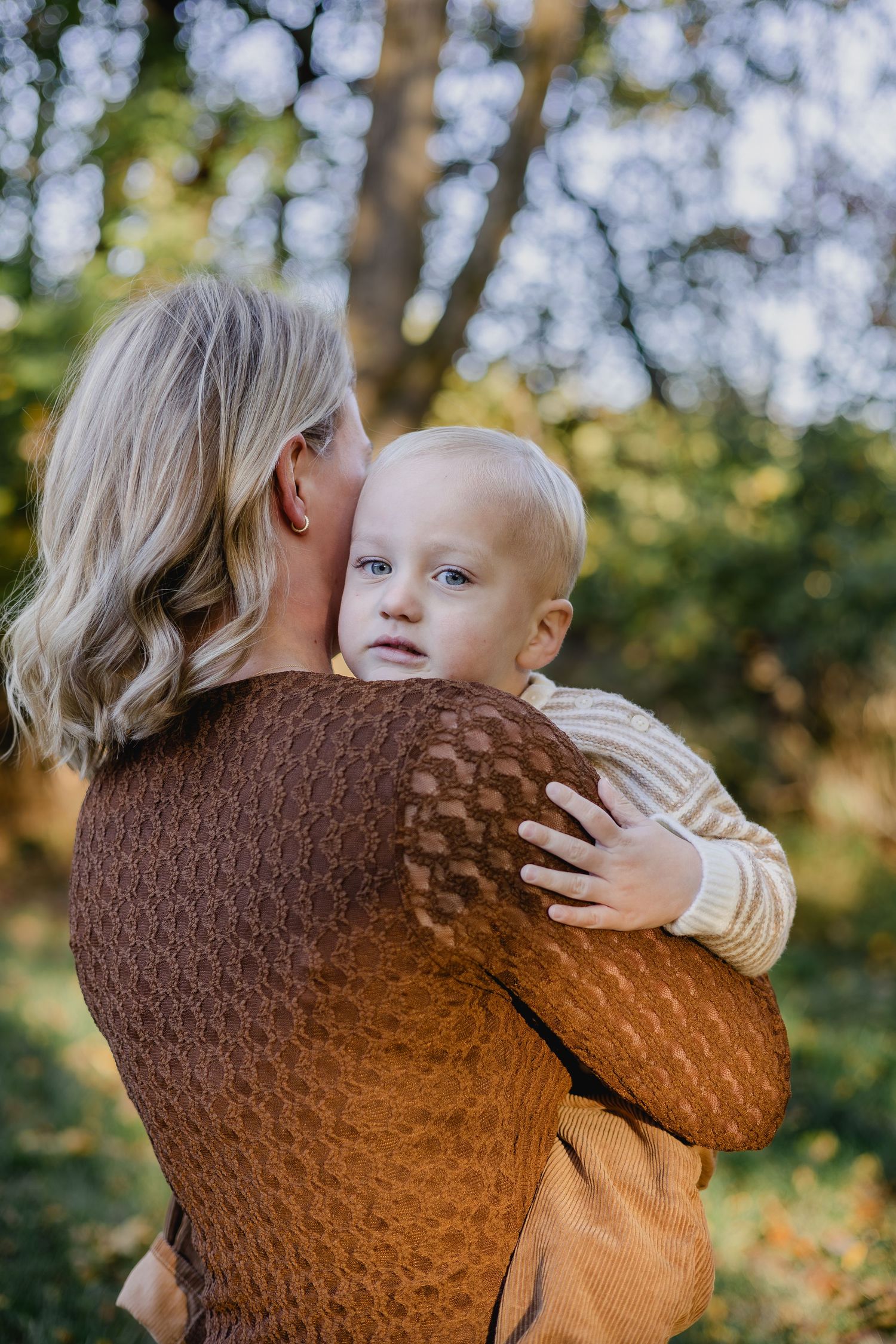 Blonde woman in brown sweater holding a blue-eyed toddler outdoors in a green park setting.