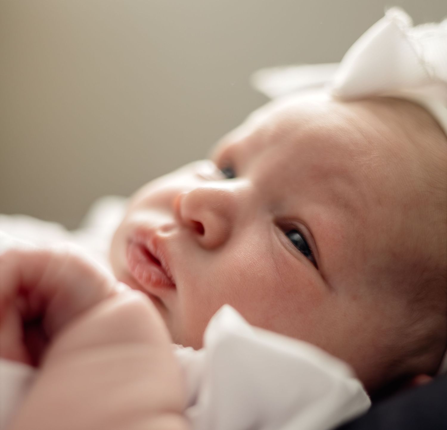 Newborn baby girl wearing a white bow headband, with delicate features and bright eyes, in a soft portrait photo.