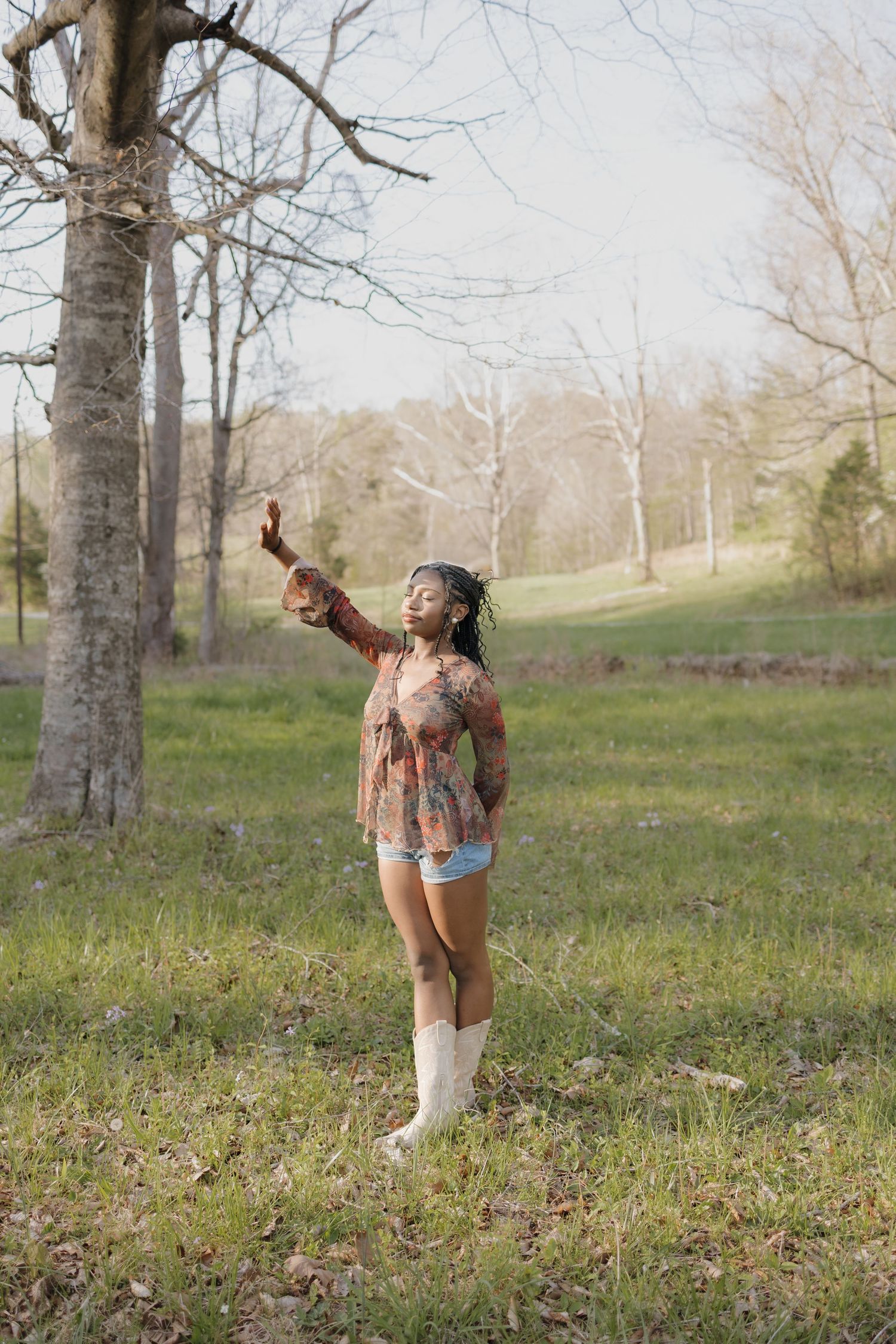 Woman in floral top and white boots reaches toward bare tree branches in a green field during early spring.