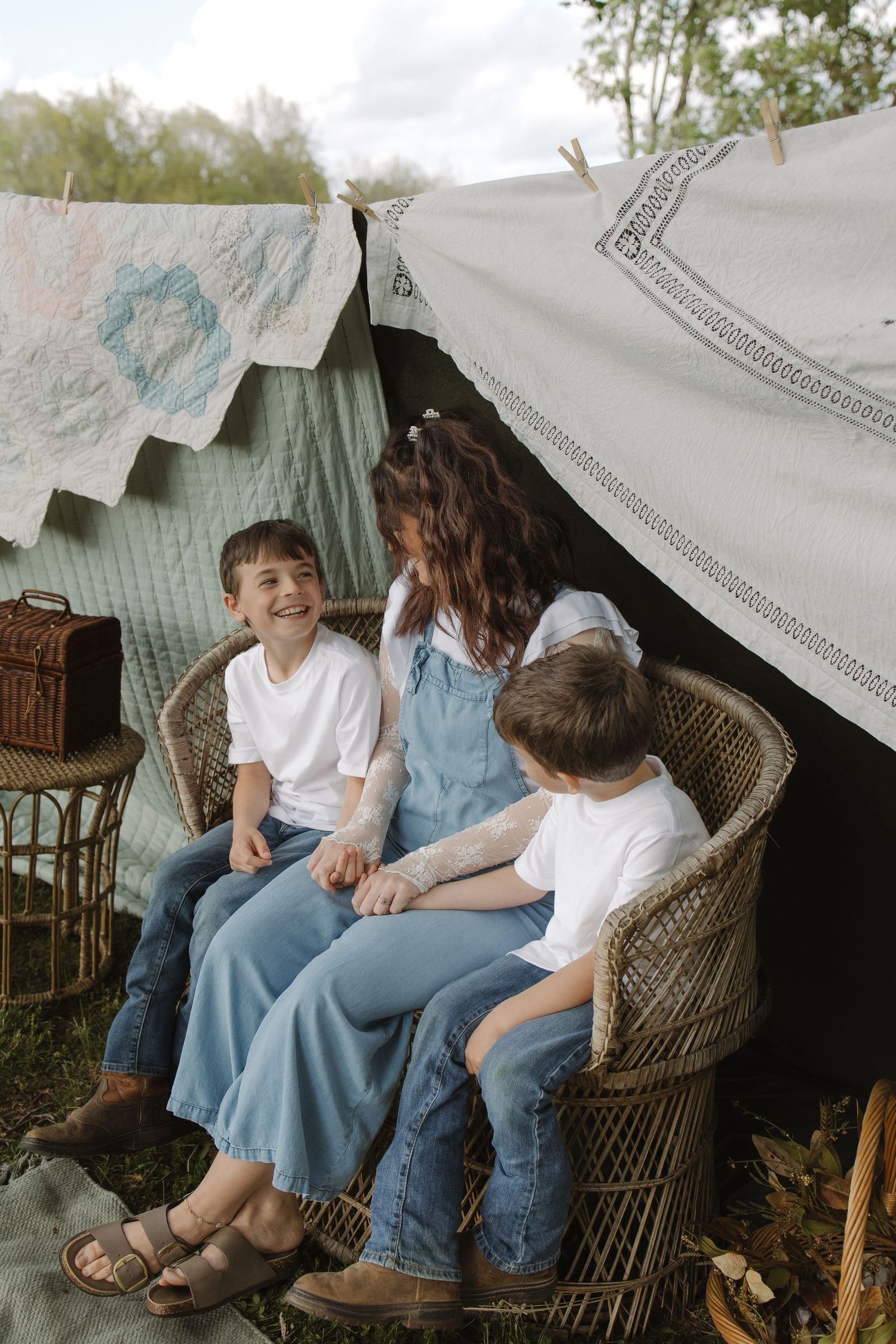A woman and two boys sit together in a wicker chair under a makeshift blanket fort outdoors.