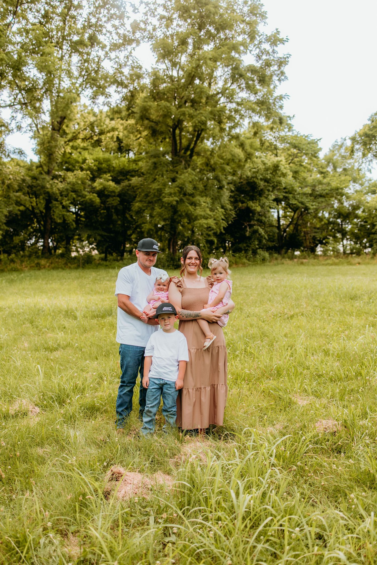 Family of five posing in a green meadow with trees, parents holding toddlers while young boy stands in front.