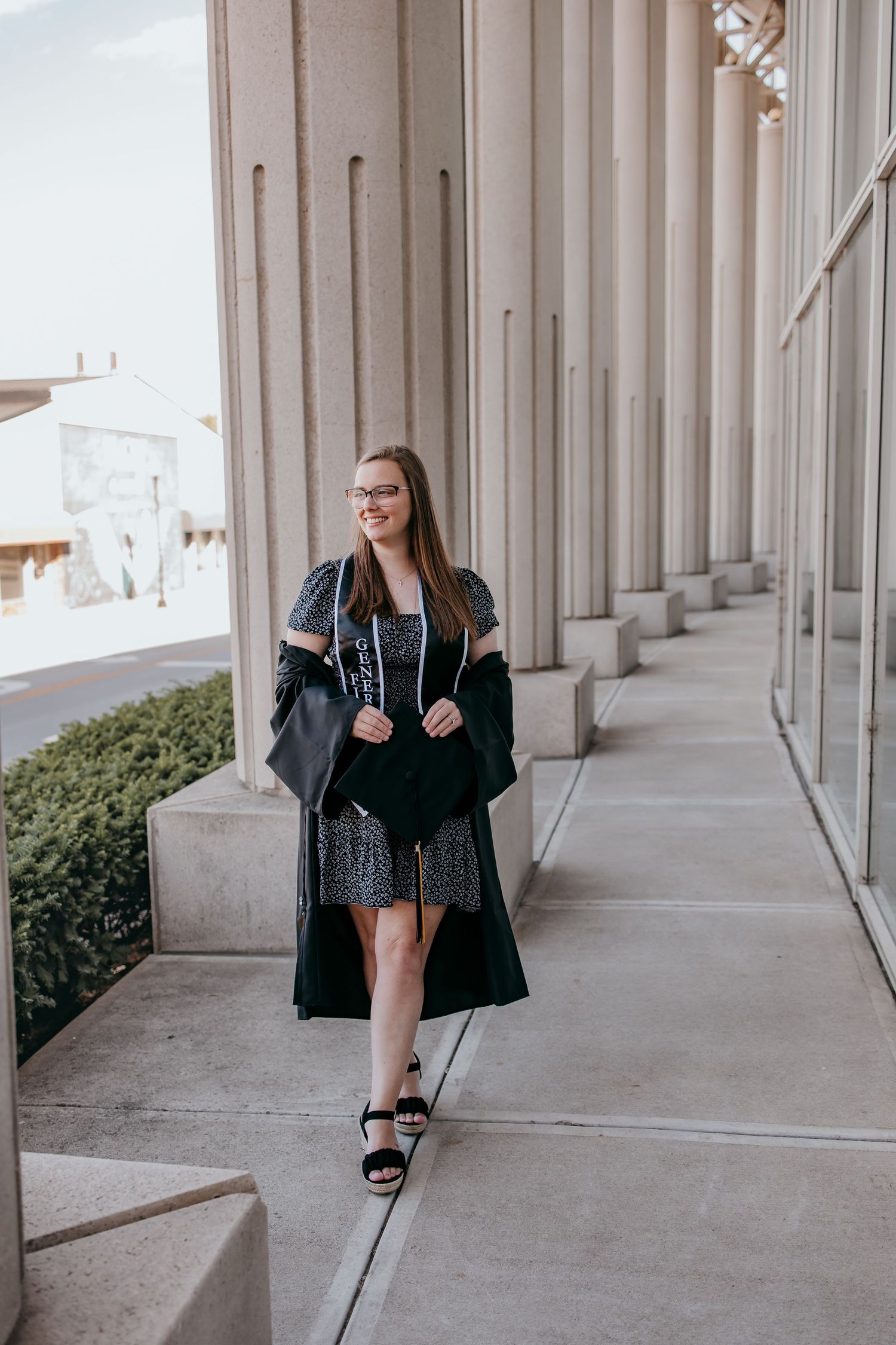 Young woman in graduation gown and patterned dress walking along columned building exterior.