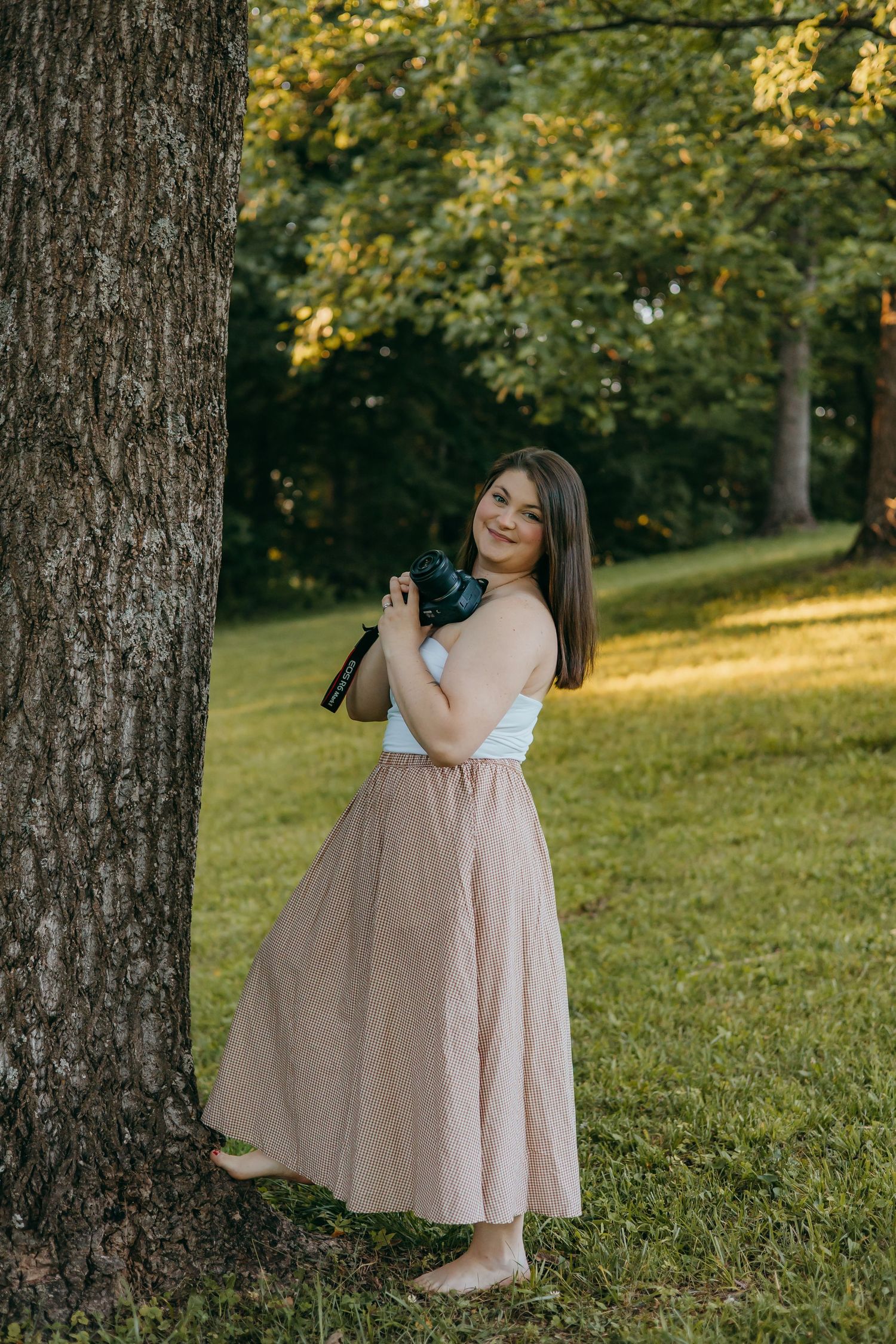 Young woman in plaid maxi skirt and white top holds camera while leaning against tree in sunny park.