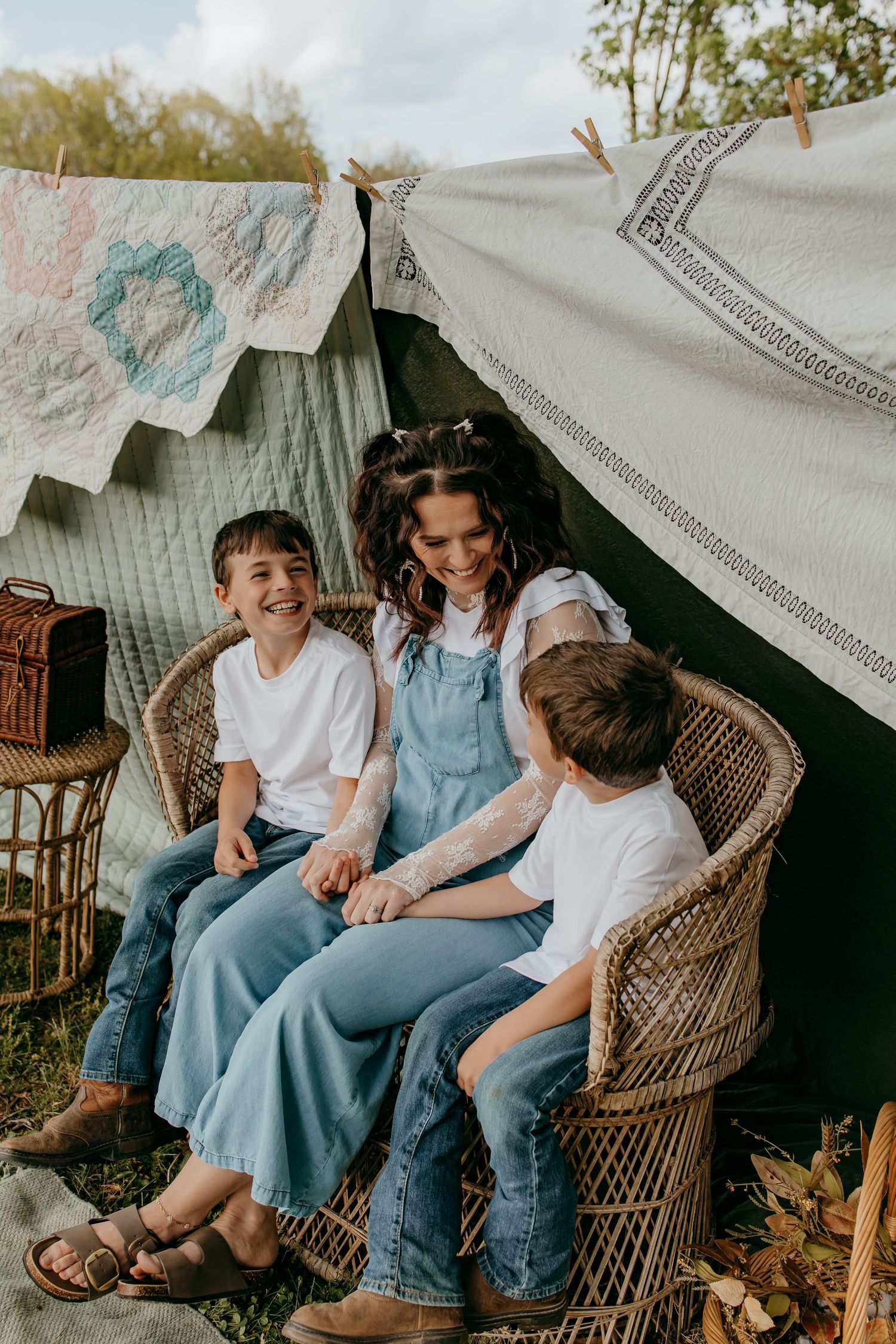 A woman in denim overalls smiles with two boys in white tees, sitting in wicker chairs under a cozy outdoor tent.
