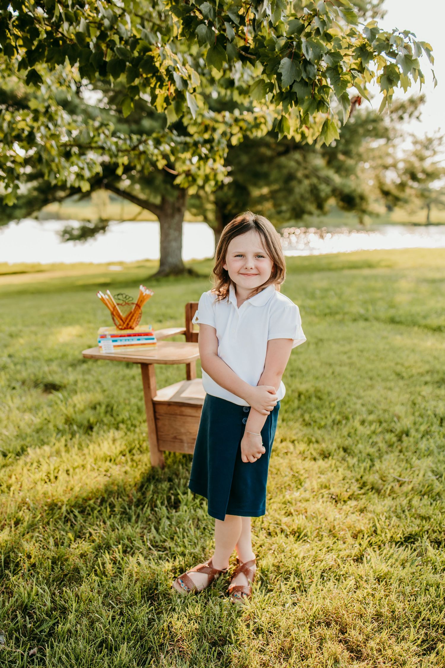 Young girl in white polo and navy skirt smiles outdoors near a wooden desk with school supplies under a tree.