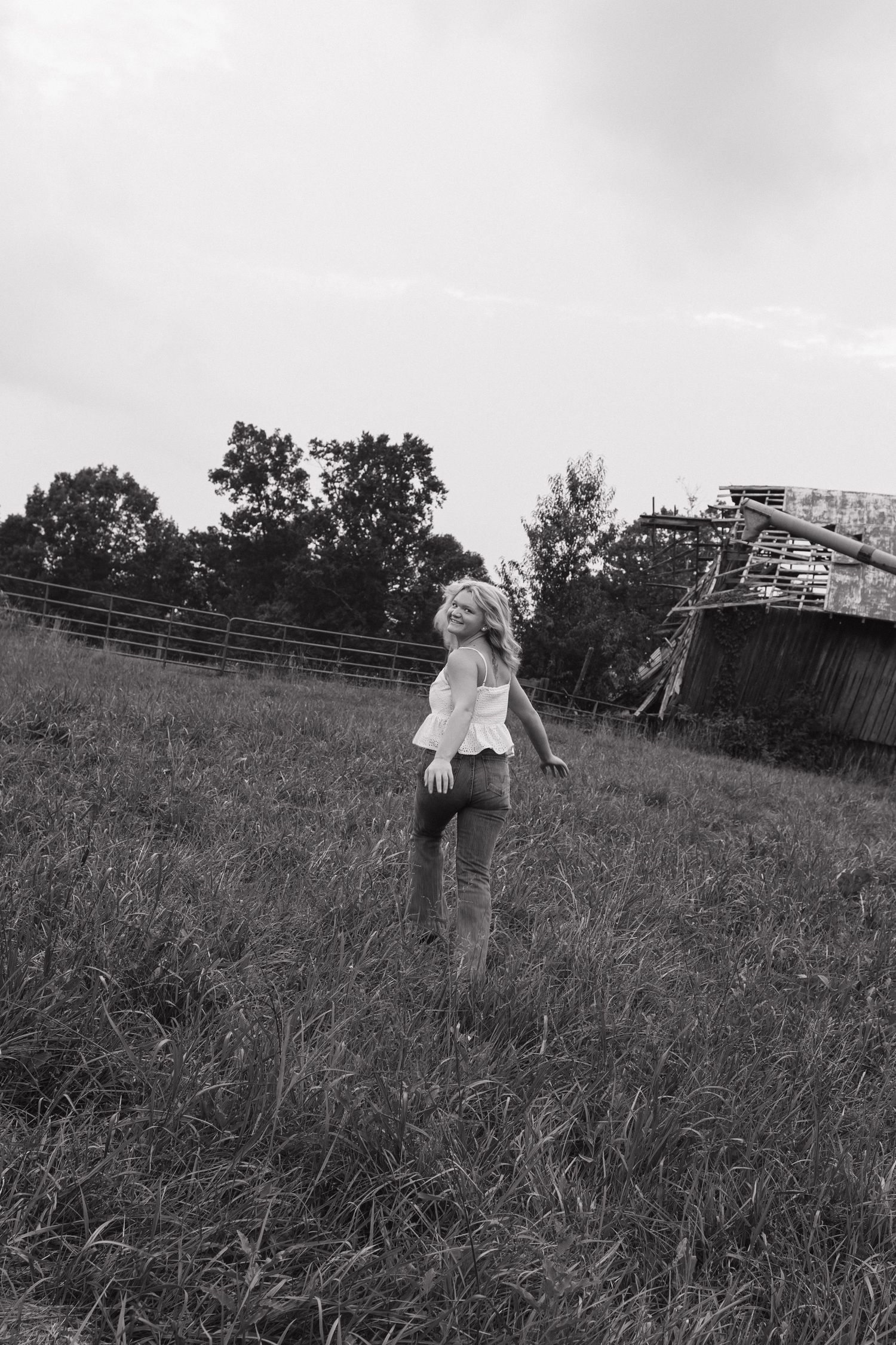 Black and white photo of a blonde woman standing in a grassy field near a rustic barn and wooden fence.