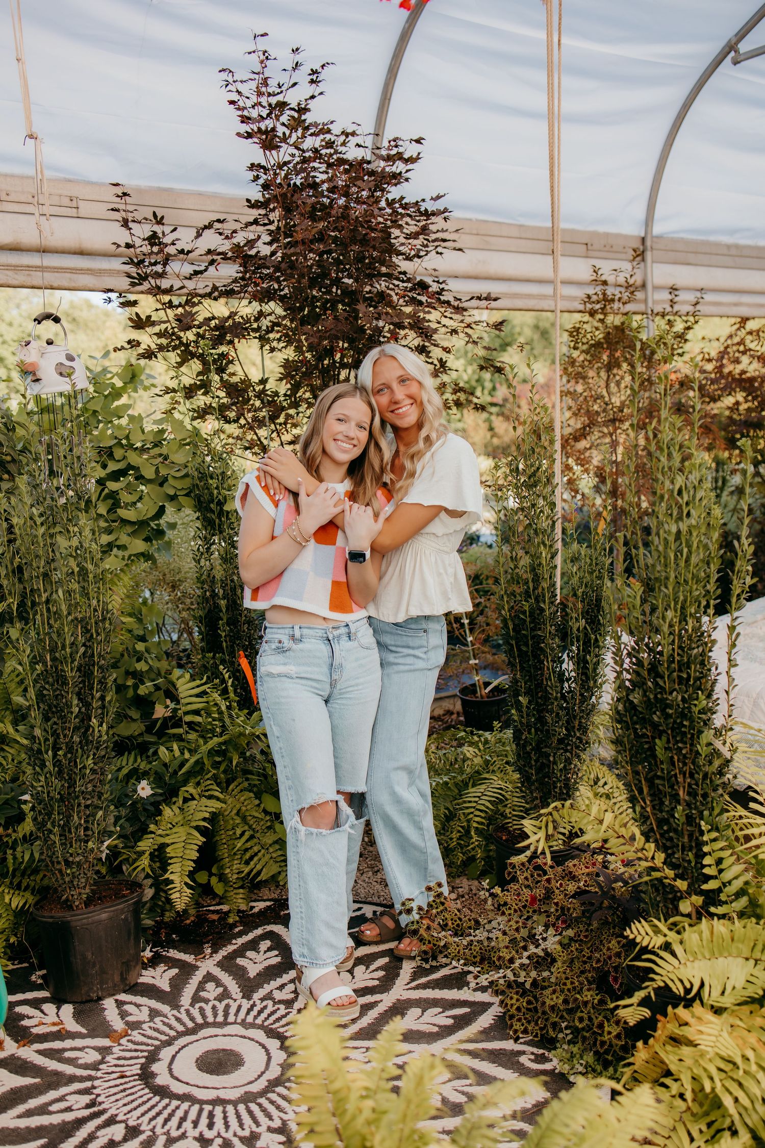 Two seniors hugging in a lush garden greenhouse surrounded by ferns and plants, wearing jeans and casual tops.