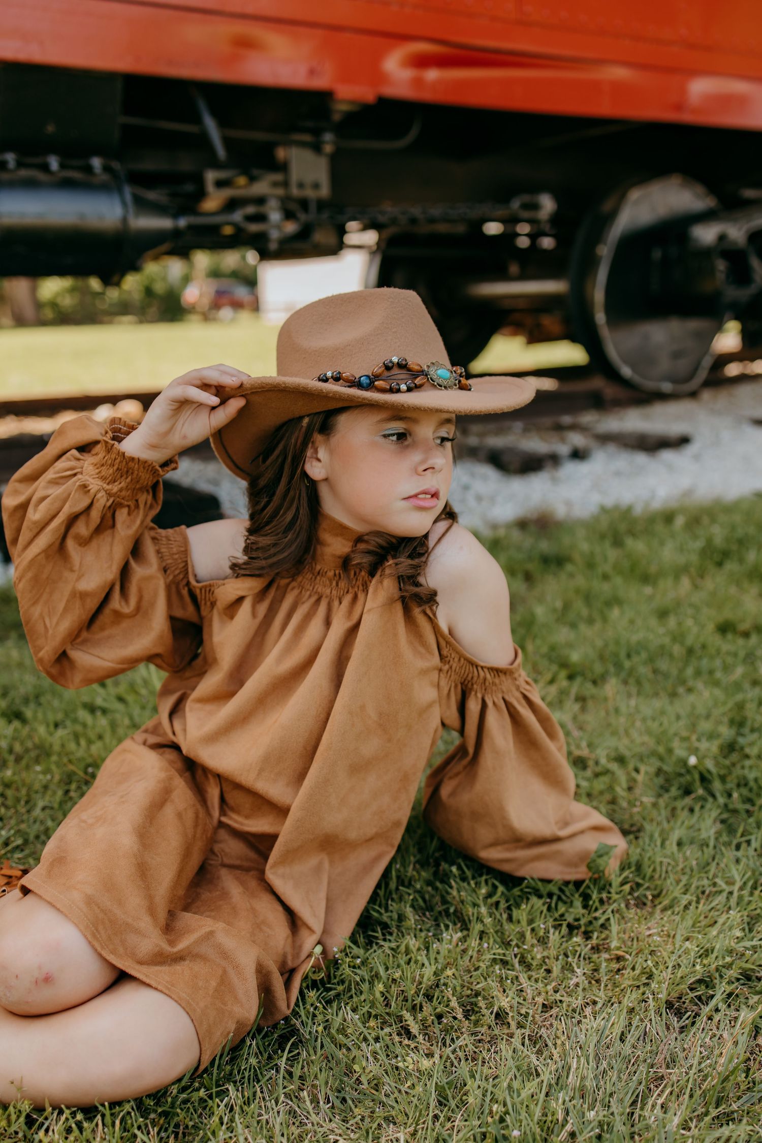 Young girl in brown suede off-shoulder dress and decorated wide-brim hat sitting on grass near red train car.