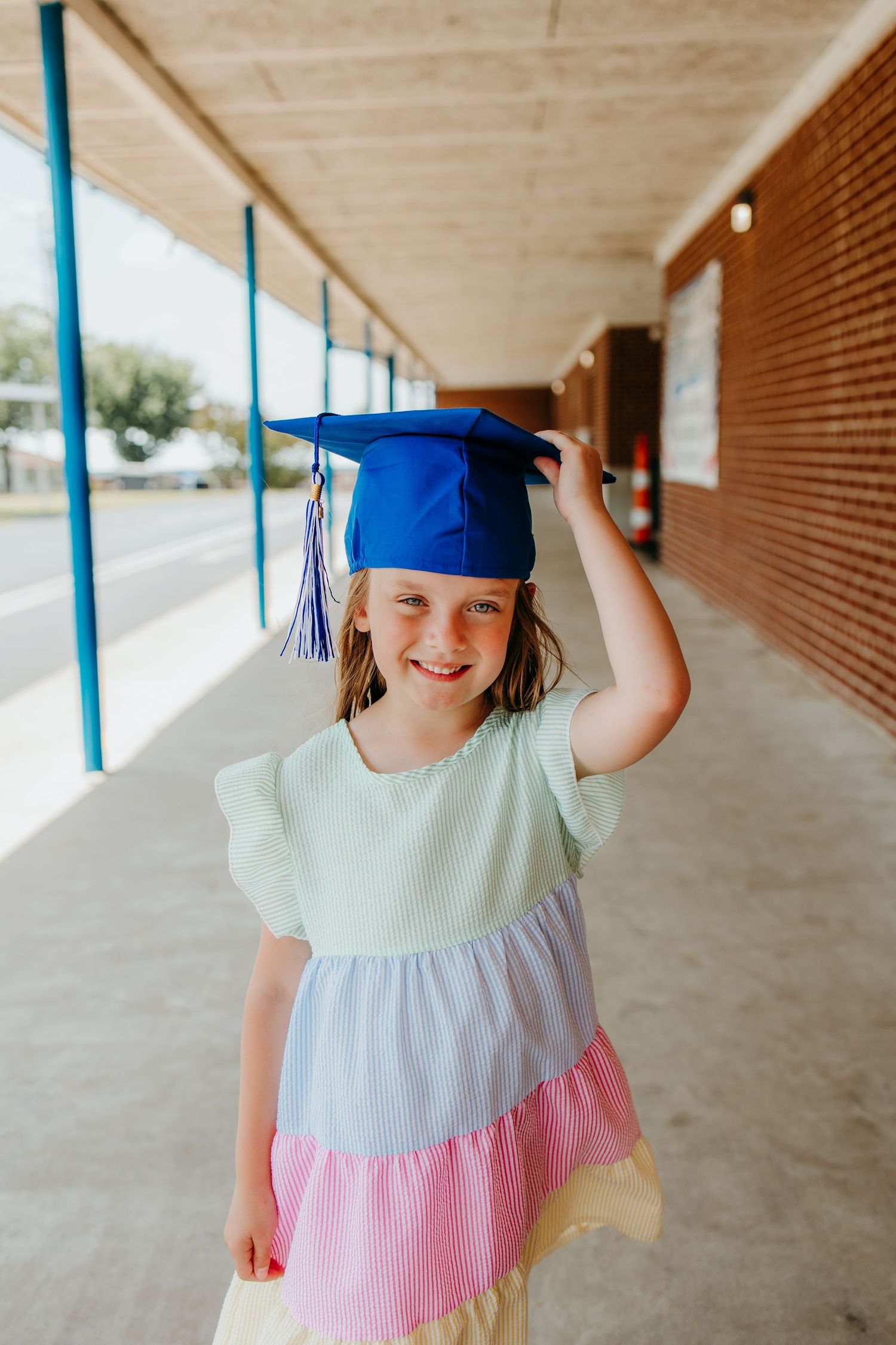 Young girl smiling in a colorful tiered dress and blue graduation cap, standing in a school hallway.