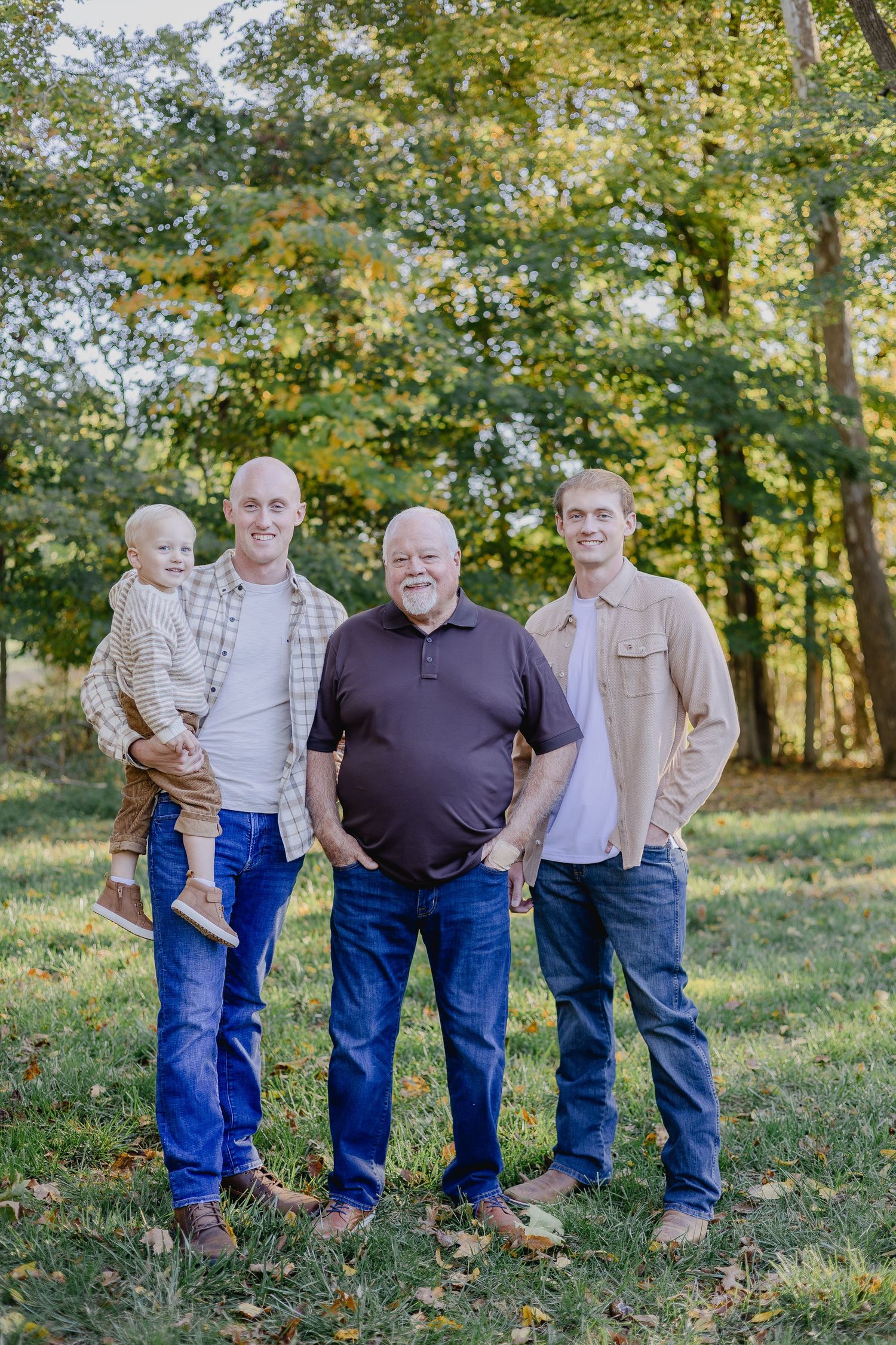 Three generations of men and a toddler pose outdoors in casual attire among autumn trees for a family portrait.