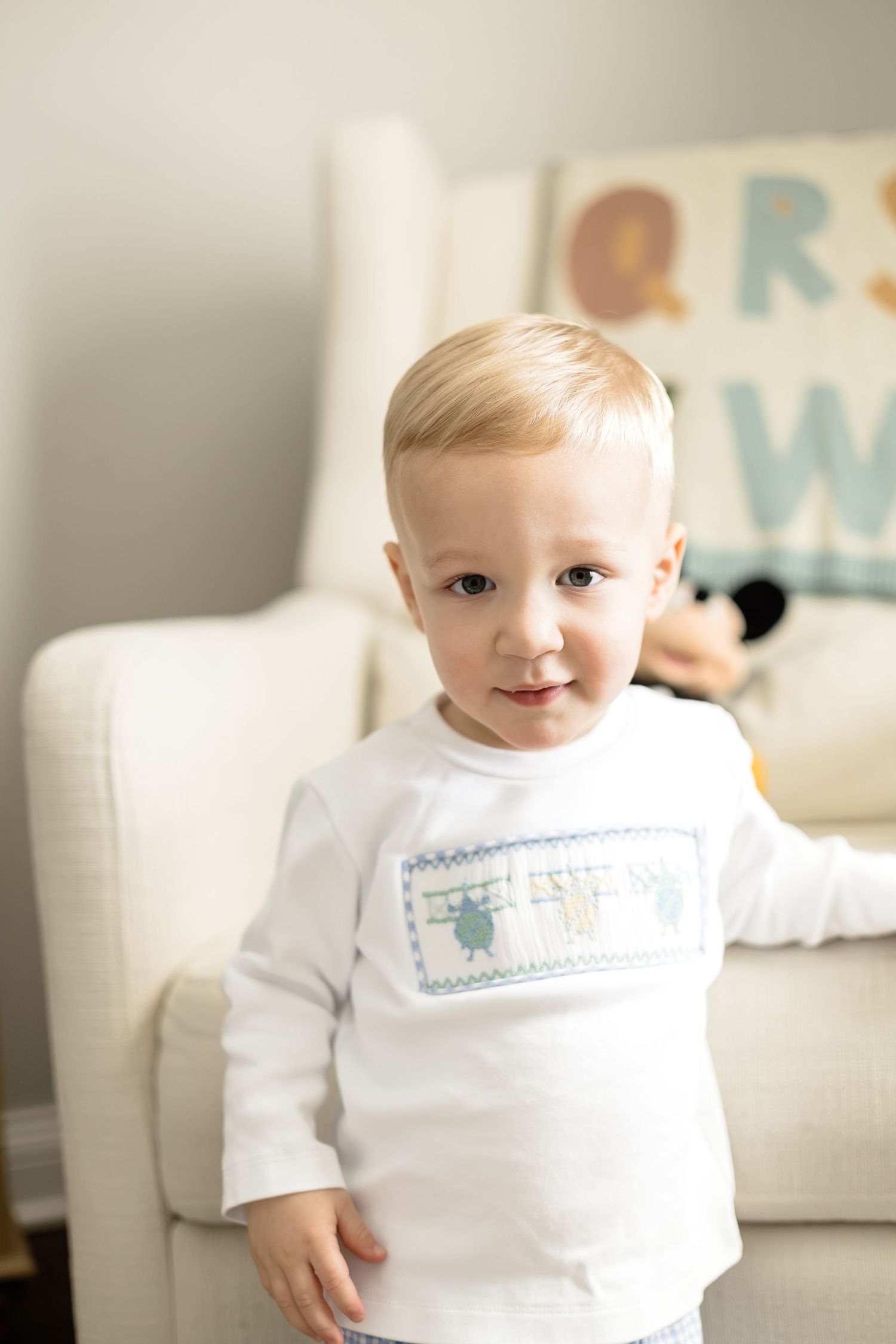Blonde toddler boy in white smocked shirt stands in front of a cream nursery chair with alphabet decor.
