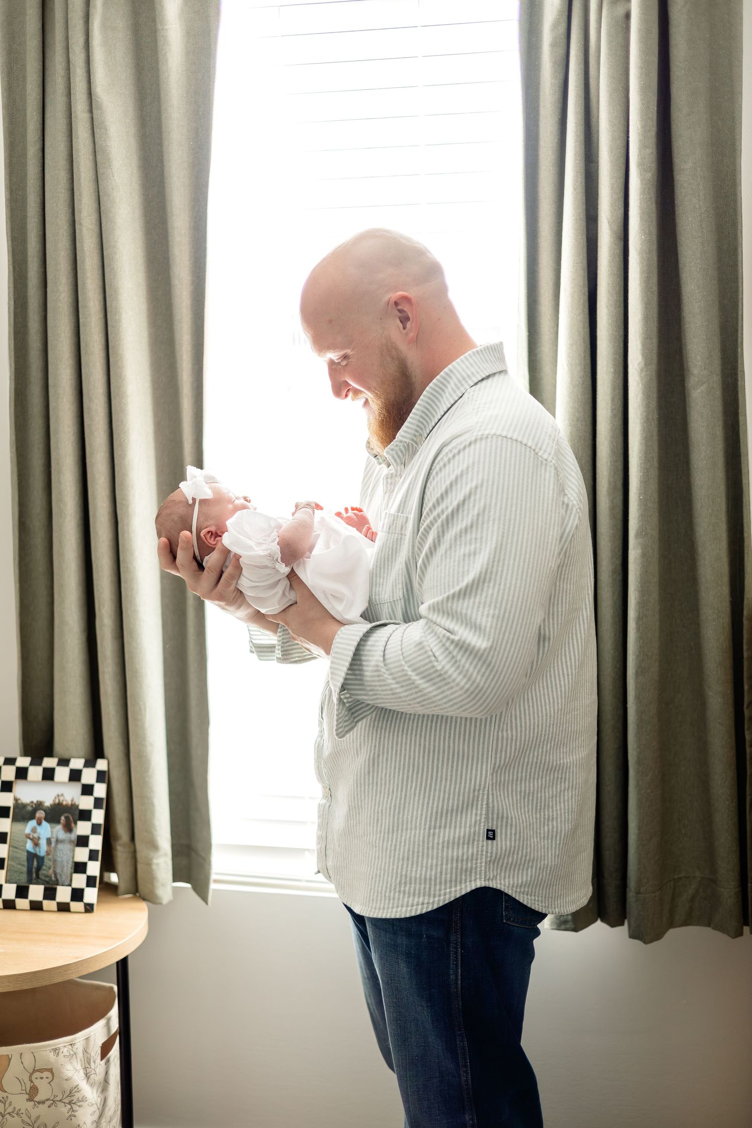 Bald father smiling at newborn baby held in his arms, standing by curtained window in soft natural light.