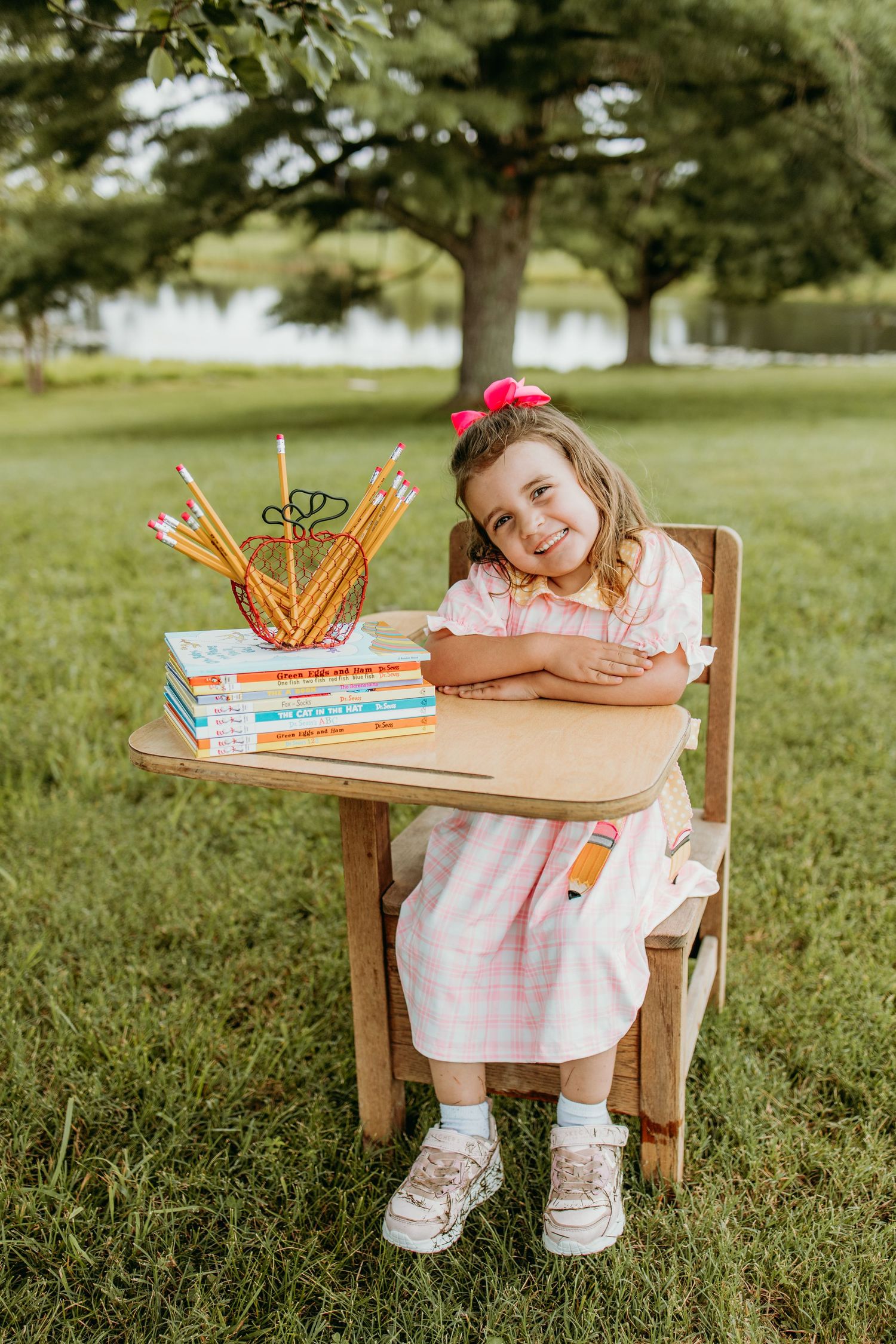 A smiling young girl in a pink dress sits at a wooden desk outdoors with books and pencils, posing for back-to-school photo.