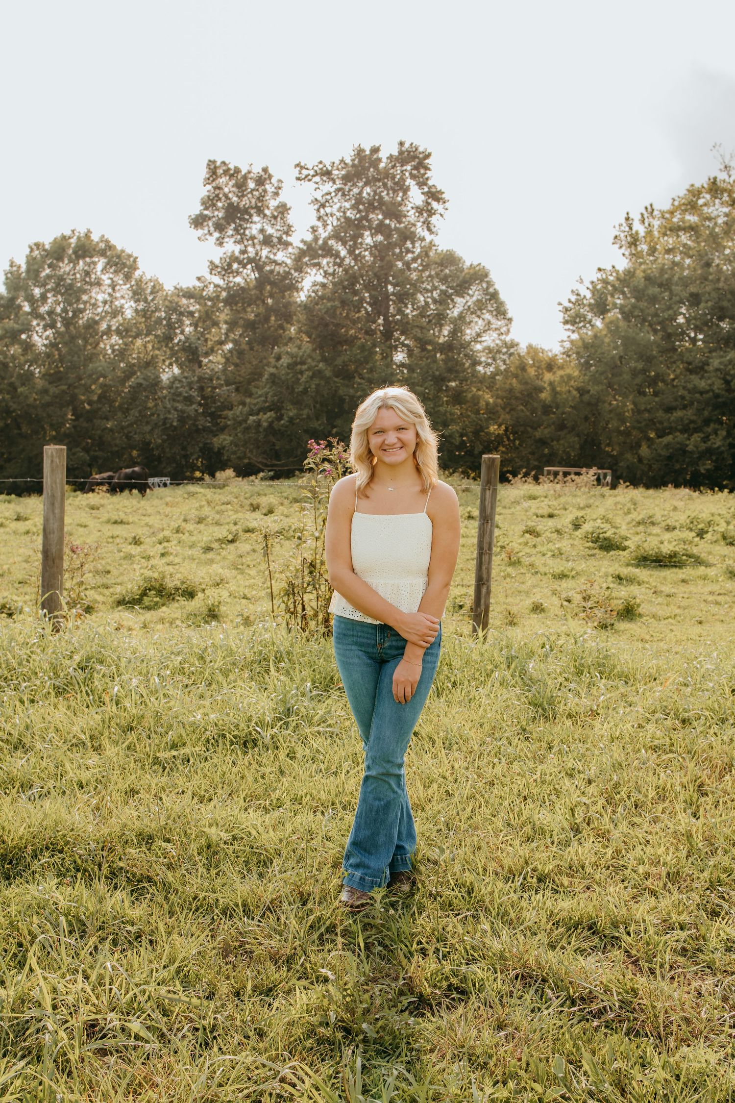 Smiling blonde woman in white tank top and flare jeans standing in a green pasture with wooden fence posts.