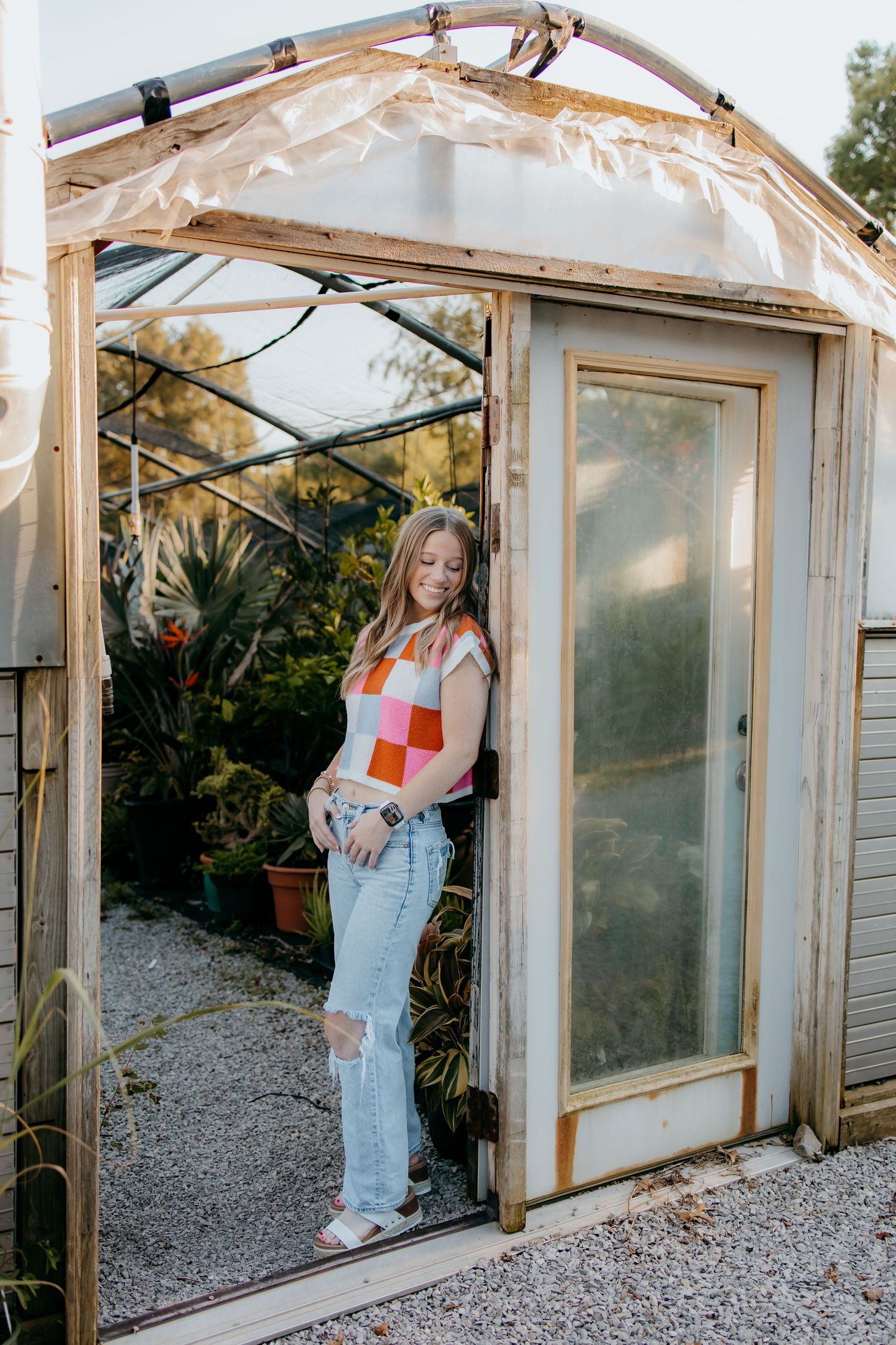 A senior in colorful checkered top and ripped jeans posing in doorway of a rustic greenhouse with plants.