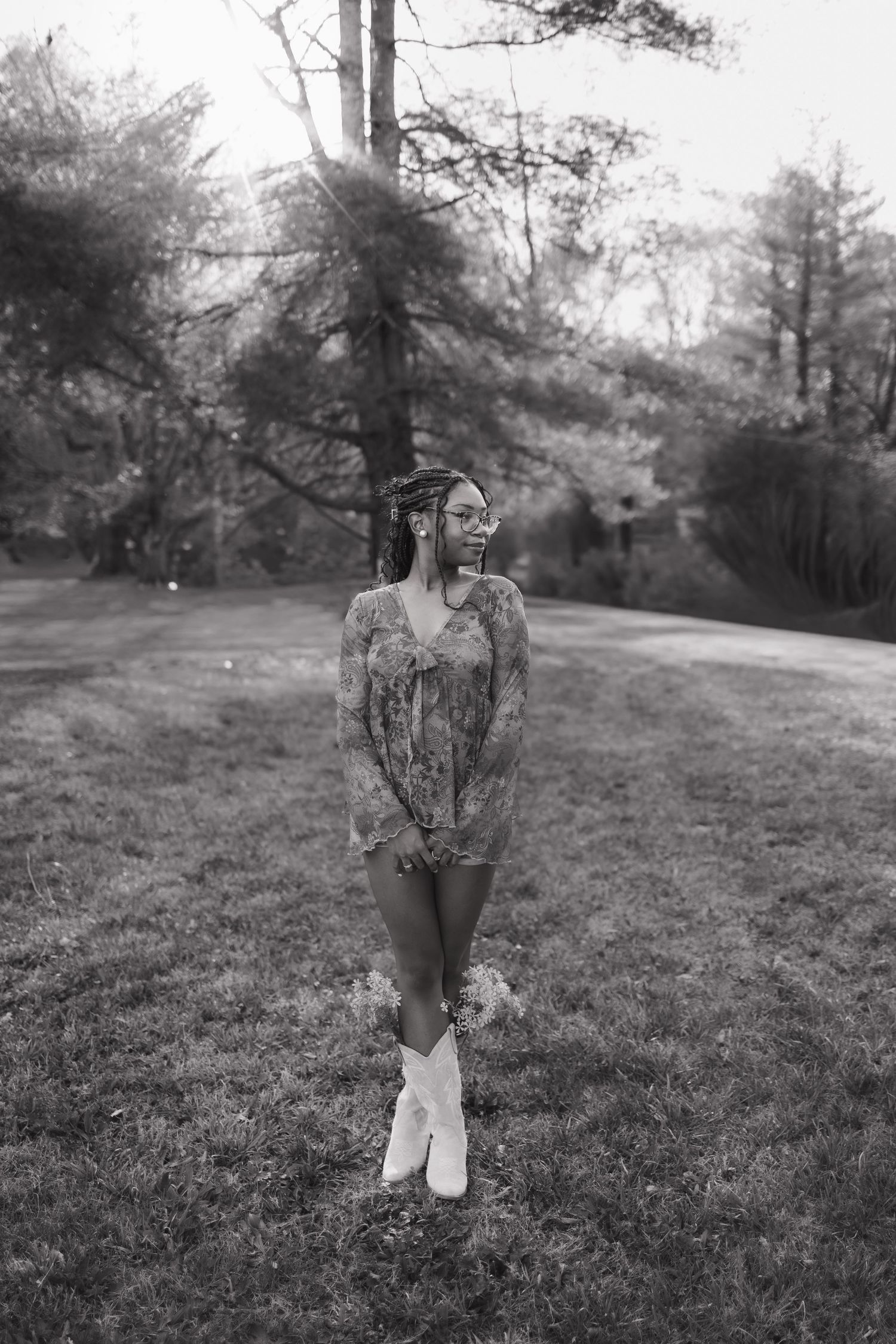 Black and white photo of a young woman in floral top, shorts, and white boots standing on grass surrounded by trees.