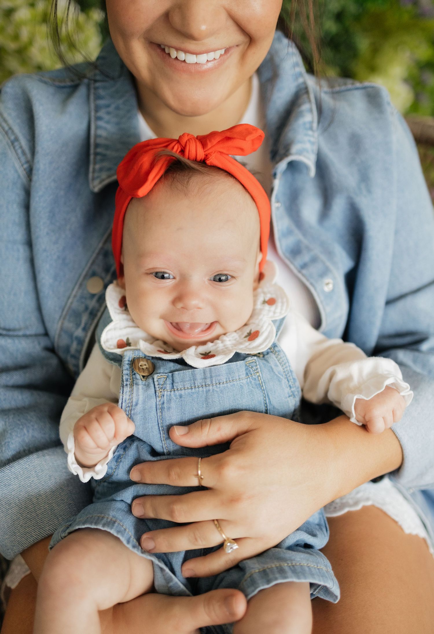 Smiling baby in denim overalls and red bow headband held by a laughing woman in a denim jacket outdoors.