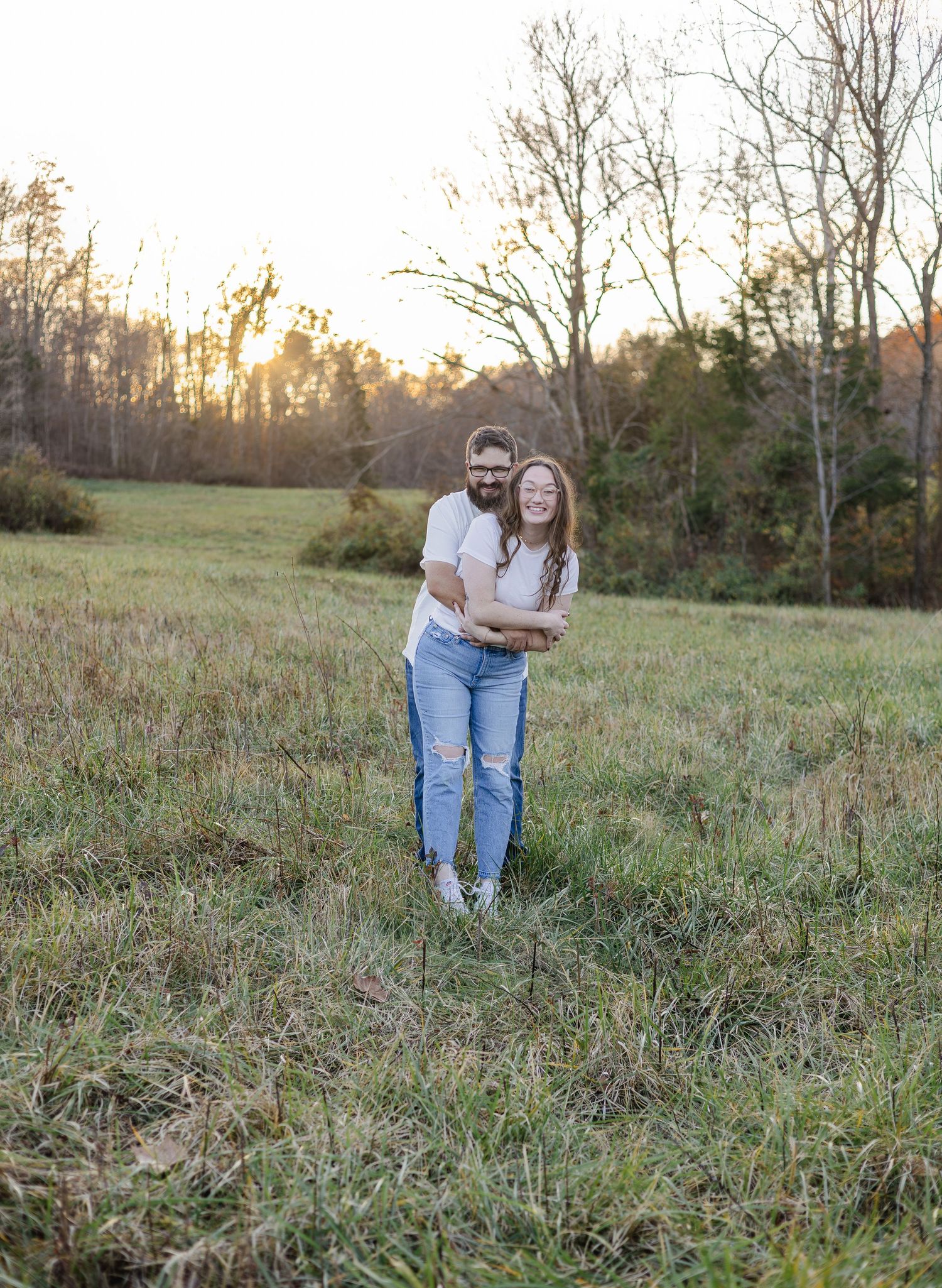 Couple embracing and smiling in a golden hour field with bare trees and warm sunset light in the background.