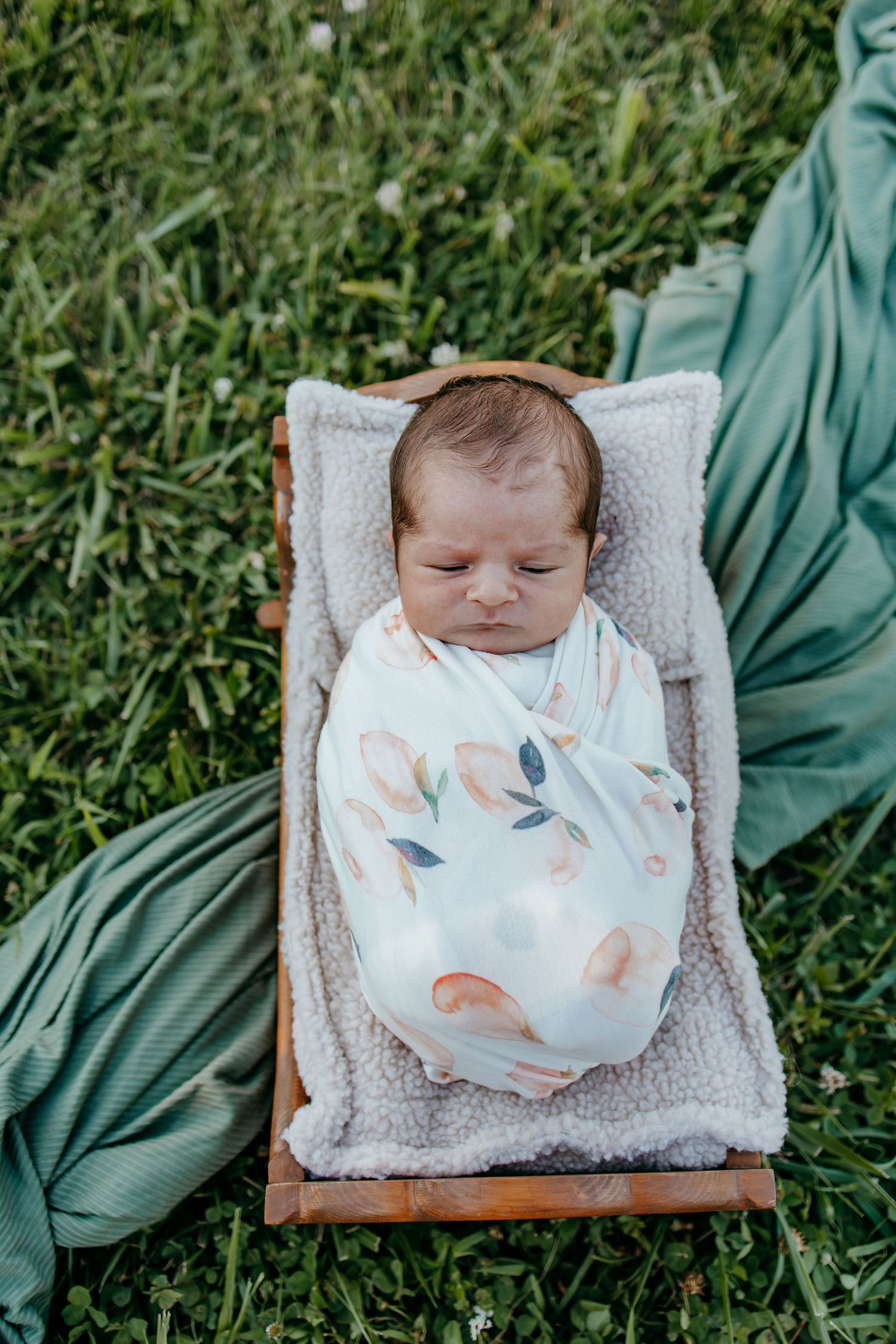 Newborn baby swaddled in floral wrap sleeping peacefully in a wooden bed on green grass outdoors.