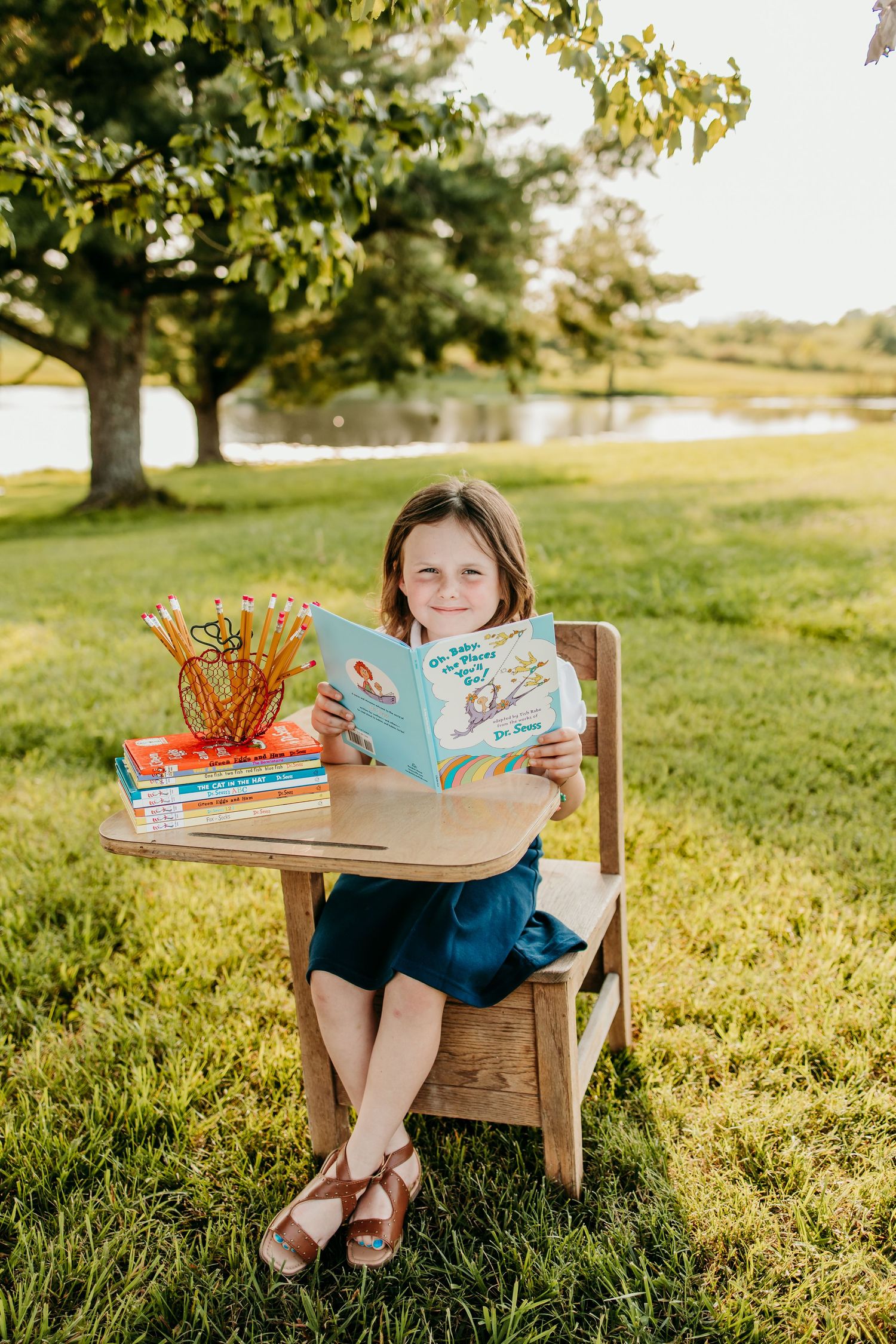 Young girl sitting at a wooden school desk outdoors reading a Dr. Seuss book, surrounded by pencils and stacked books.
