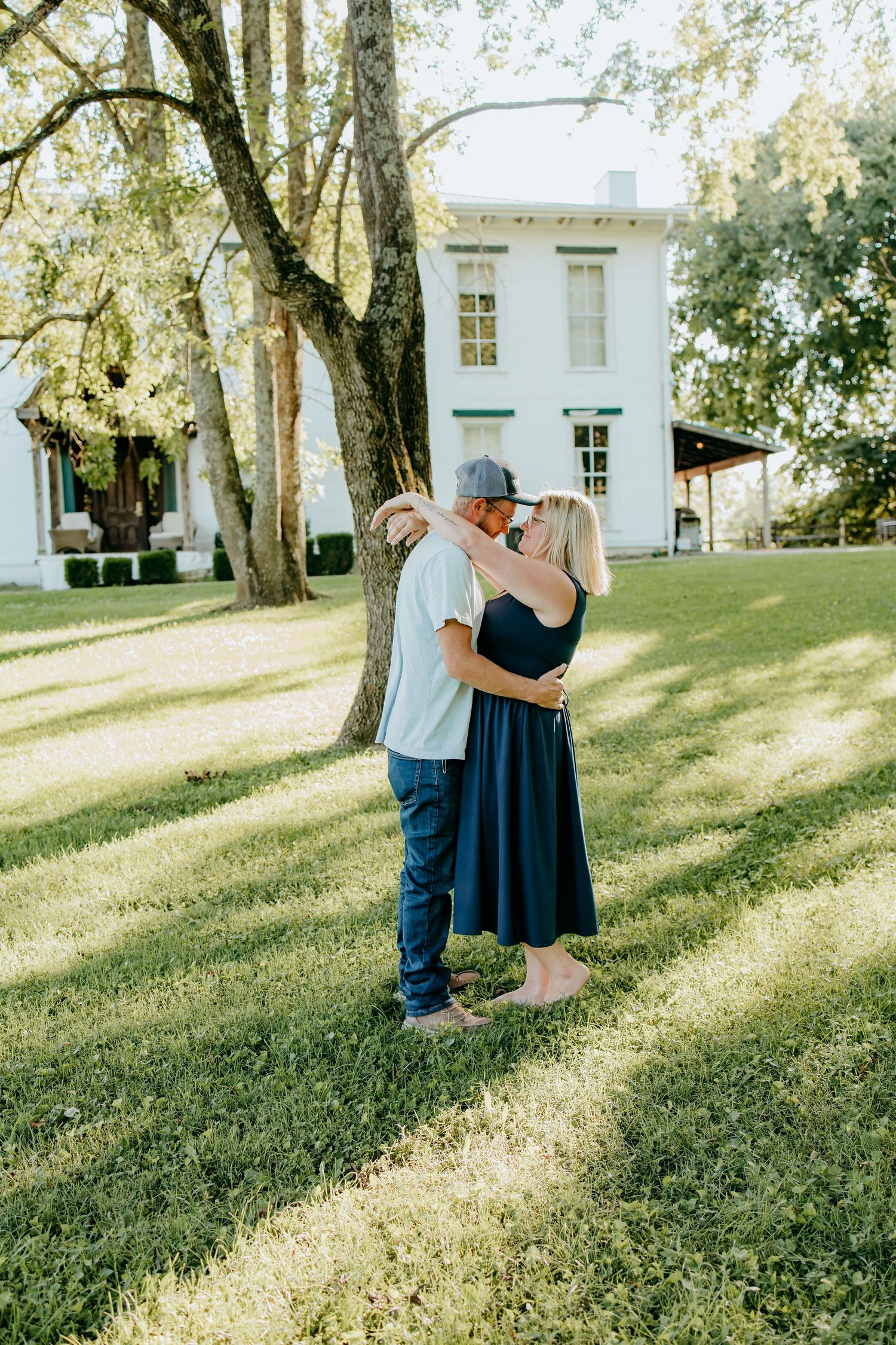 Couple sharing a romantic embrace on green lawn with white historic house and large tree in background.