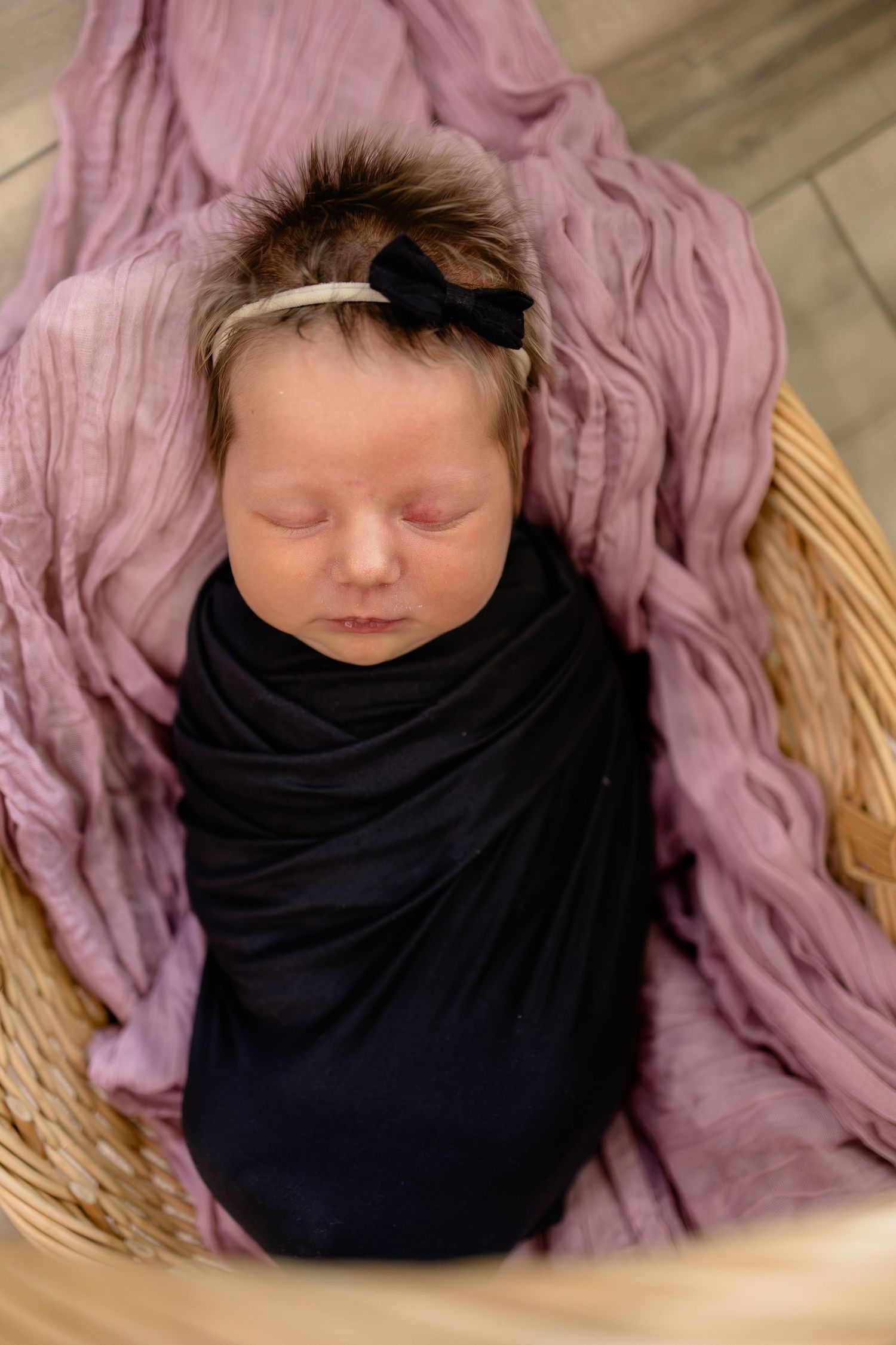 Newborn baby swaddled in black wrap with purple blanket and bow headband sleeping peacefully in wicker basket.