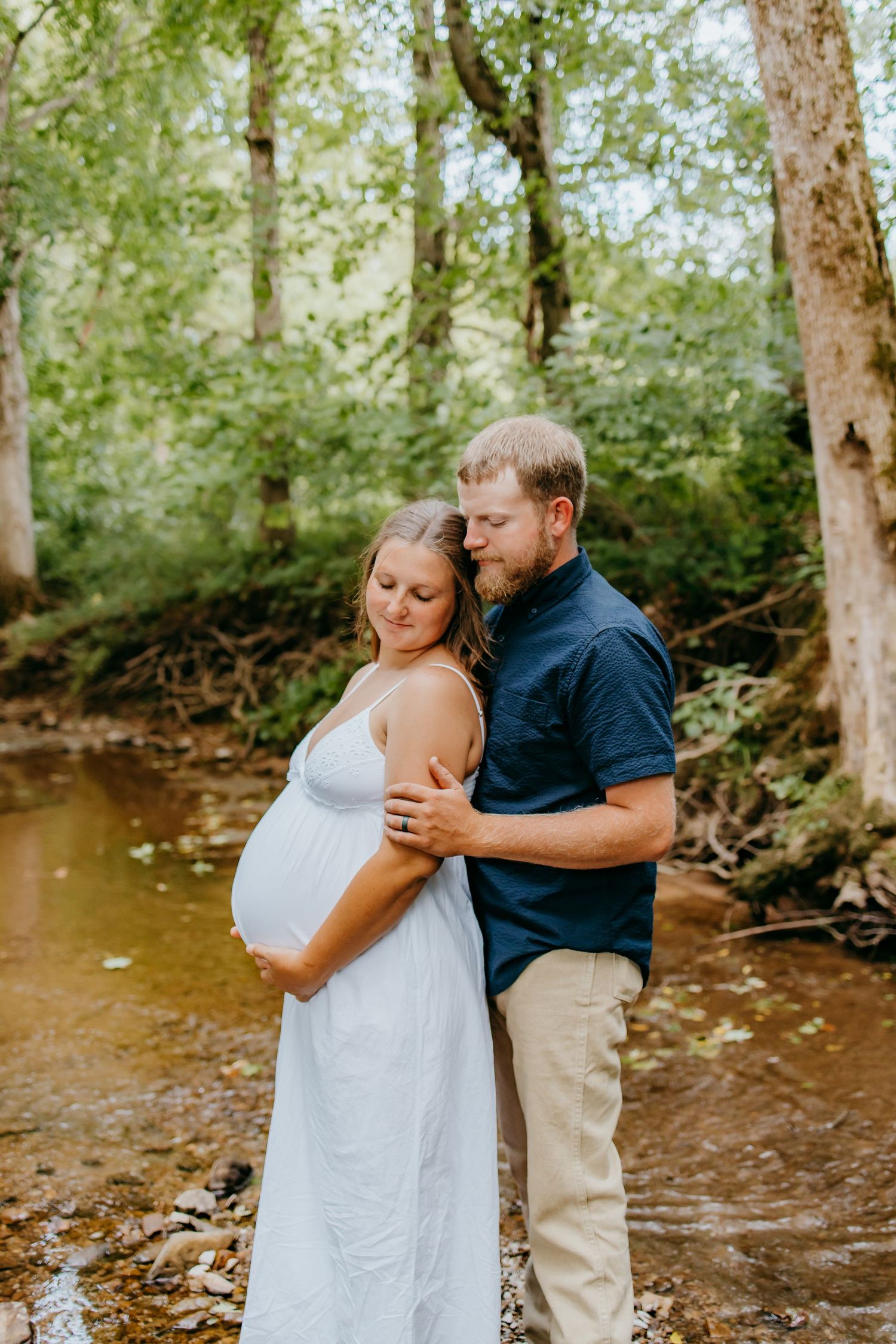 Couple maternity photo in forest creek, pregnant woman in white dress embraced by man in navy shirt.