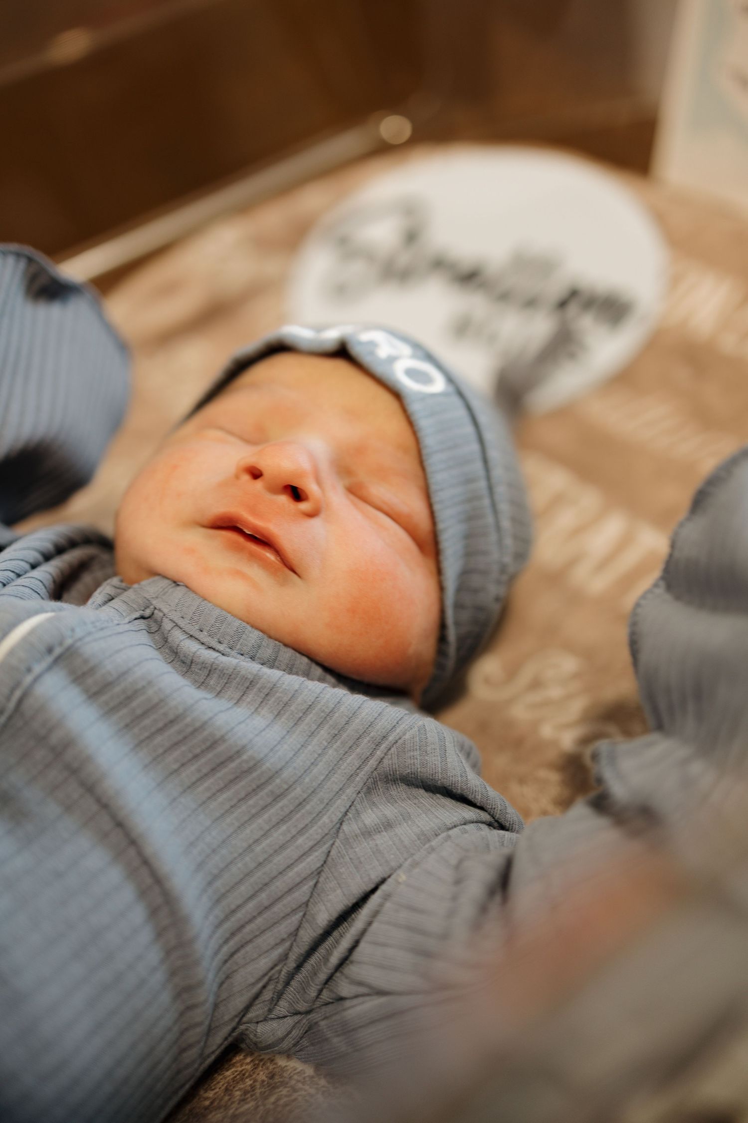 Newborn baby in gray outfit and knit hat lying peacefully in a wooden box with a name sign.
