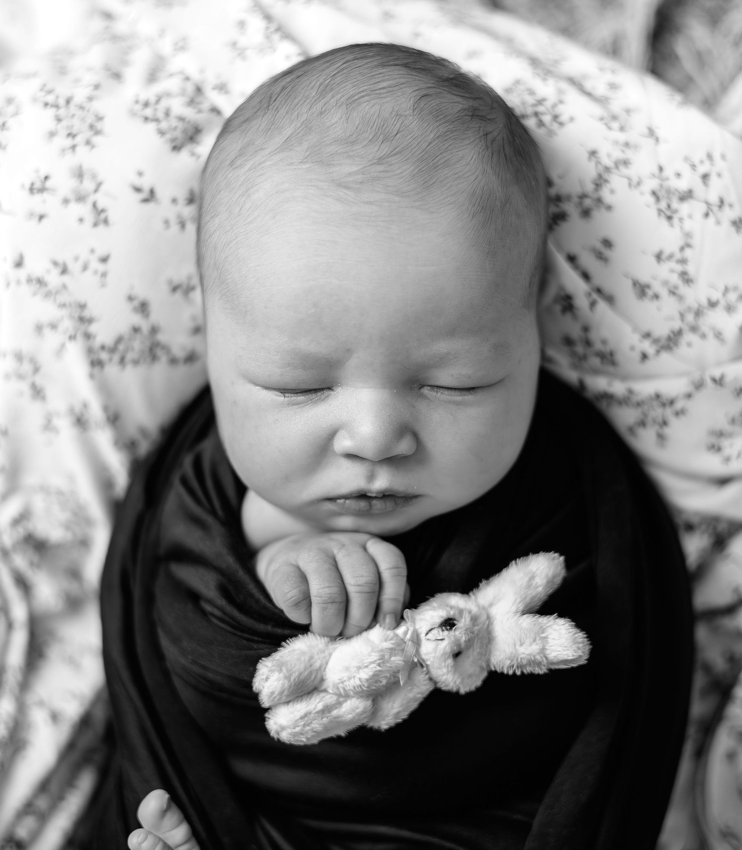 Black and white photo of a sleeping newborn baby swaddled in dark wrap, holding a small white stuffed bunny toy.