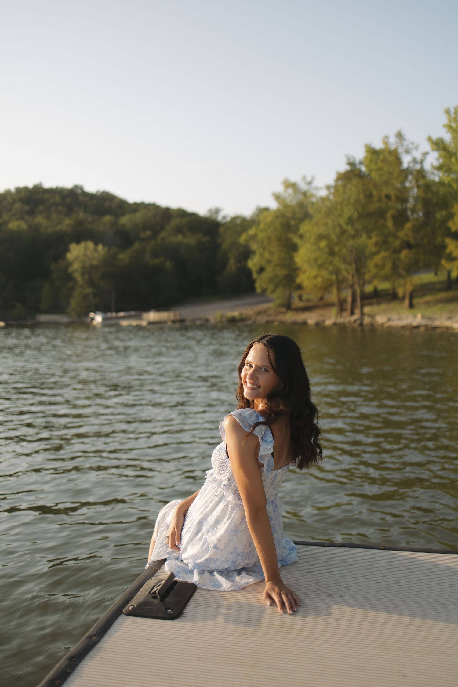 Young woman in white dress sitting on a dock by a lake, smiling, with lush green trees in the background.
