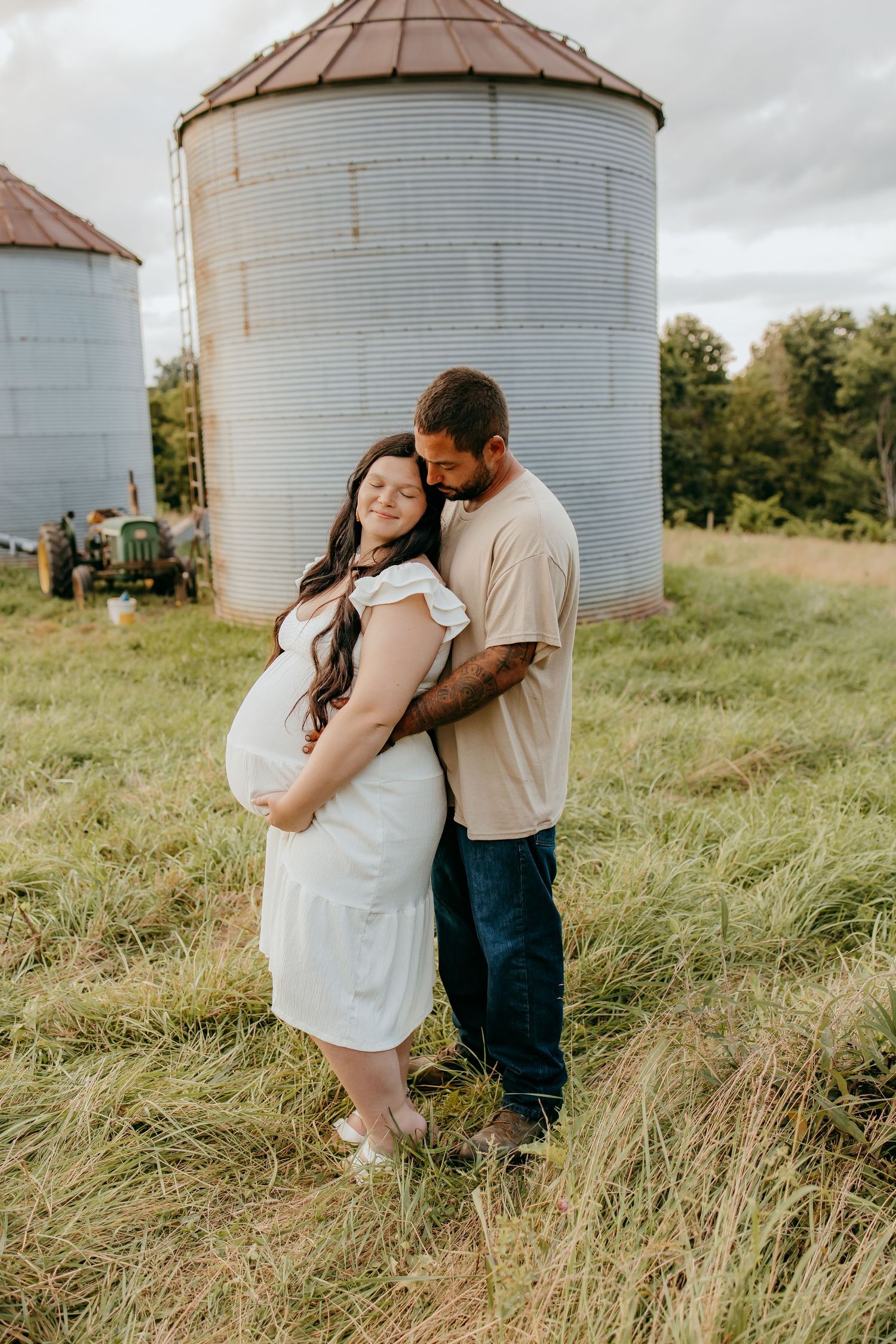 Pregnant couple embracing in a farm field with grain silos, woman in white dress, man in tan shirt and jeans.