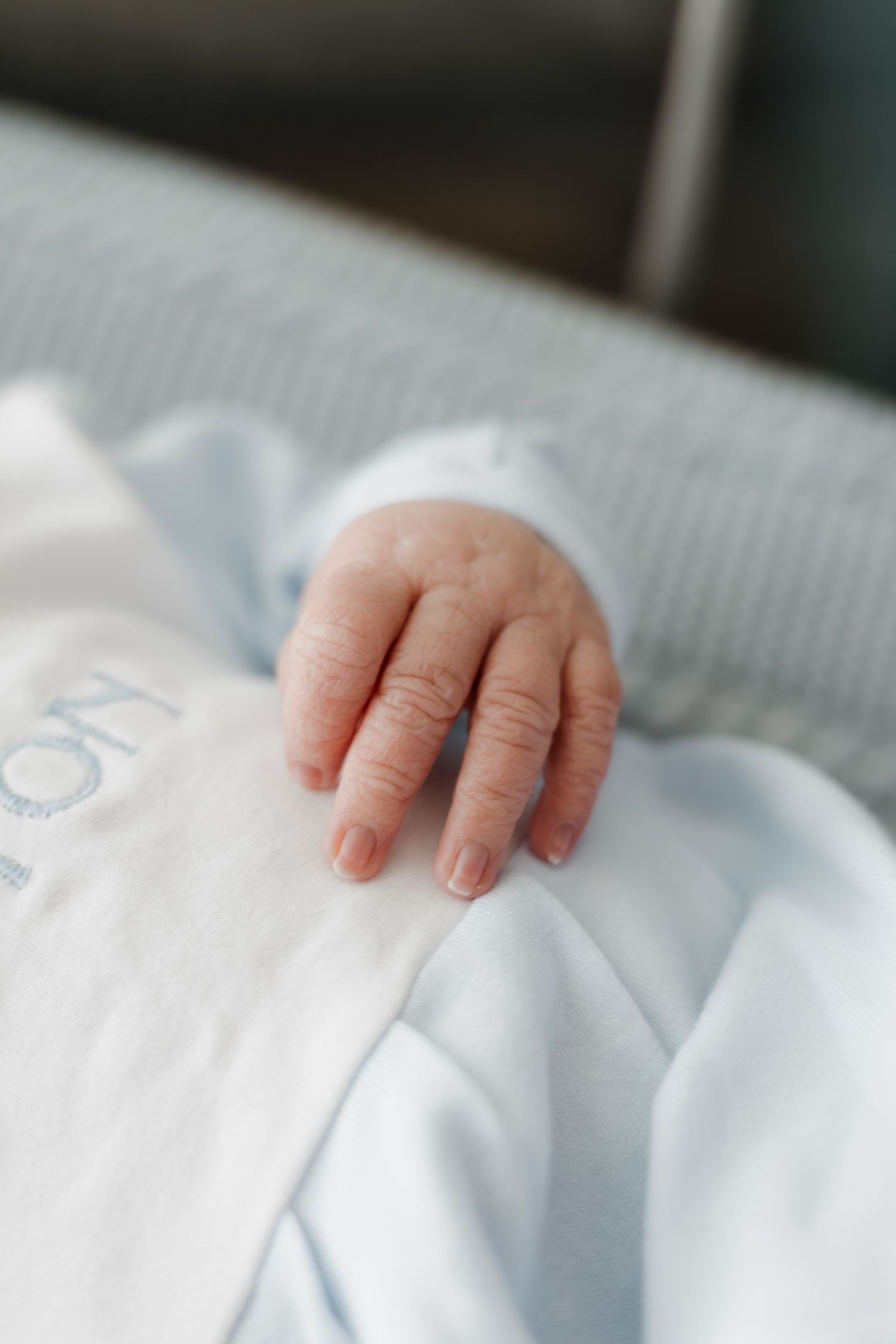 Close-up of a newborn baby's tiny hand resting on a white onesie in a hospital bassinet.