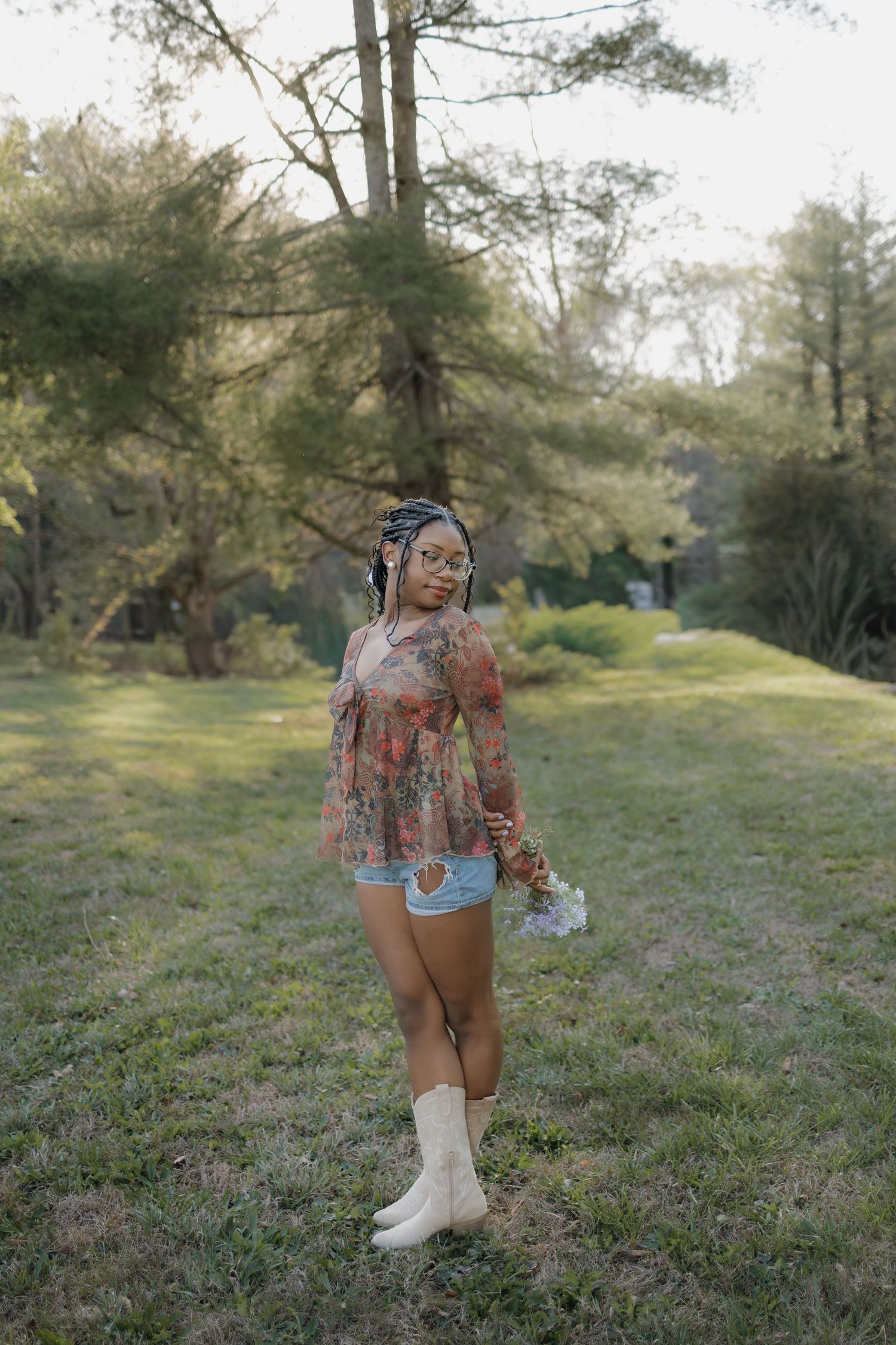 Young Black woman in floral top, denim shorts, and white boots standing in a lush green park holding flowers.