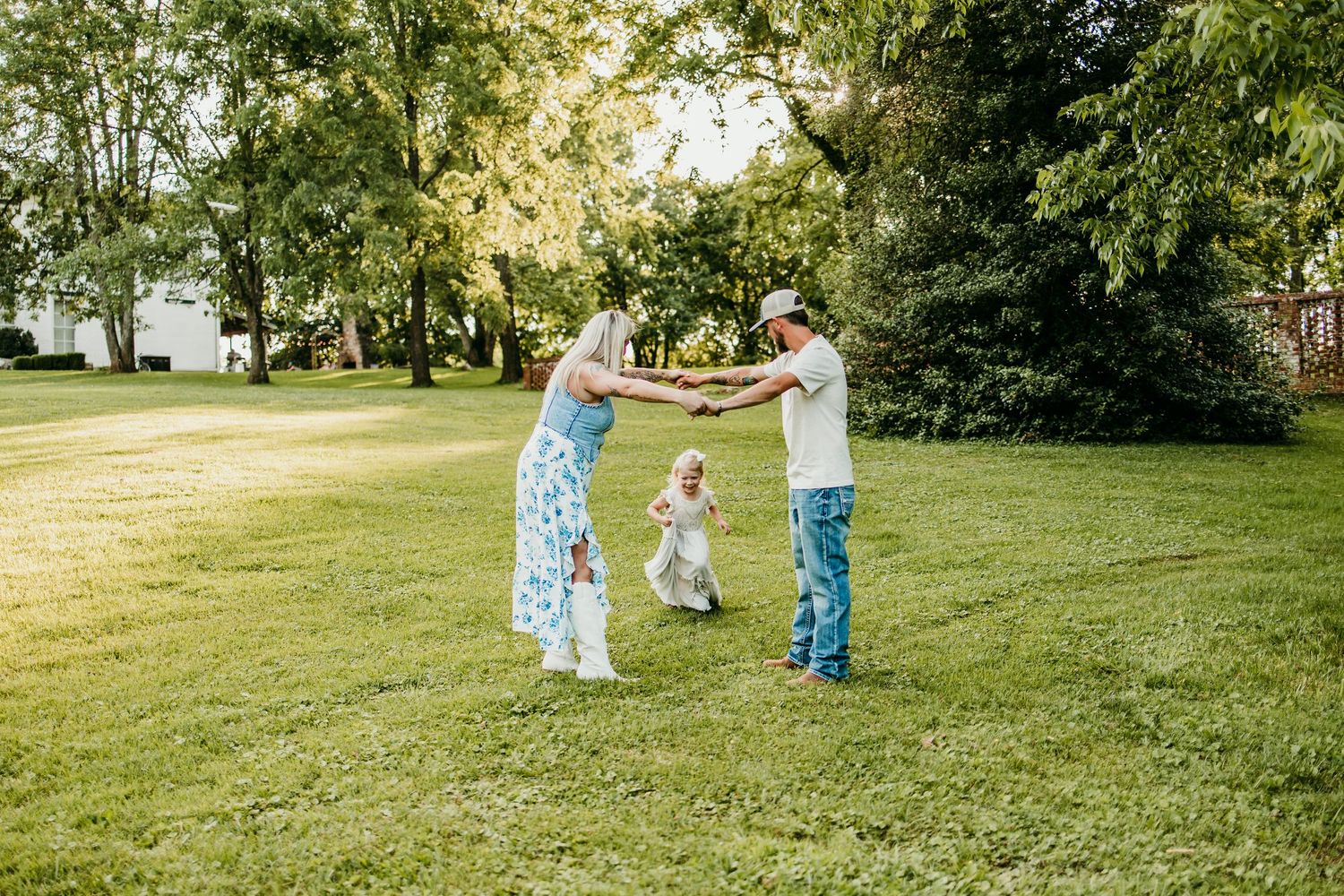 A family of three holds hands and dances together on a sunny green lawn surrounded by lush trees.