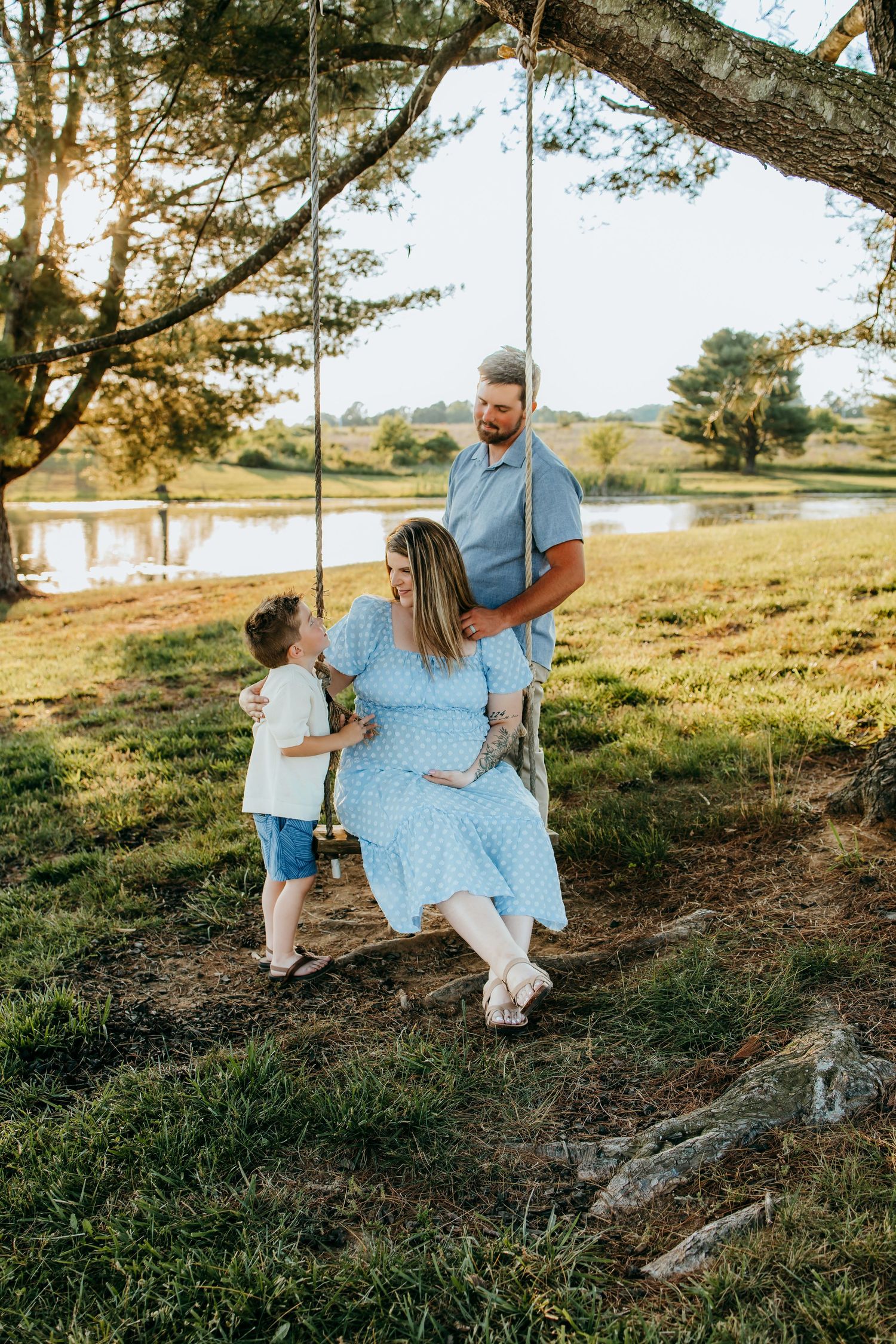 Pregnant woman in blue dress sits on tree swing with toddler and husband in sunny rural setting near pond.