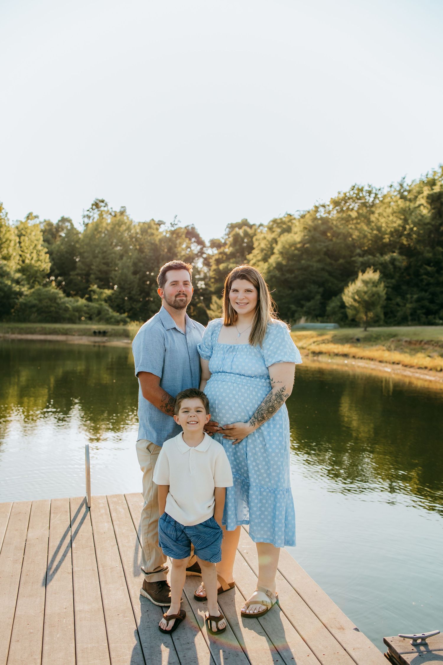 Pregnant woman in blue dress with husband and son posing on a wooden dock by a serene lake at sunset.
