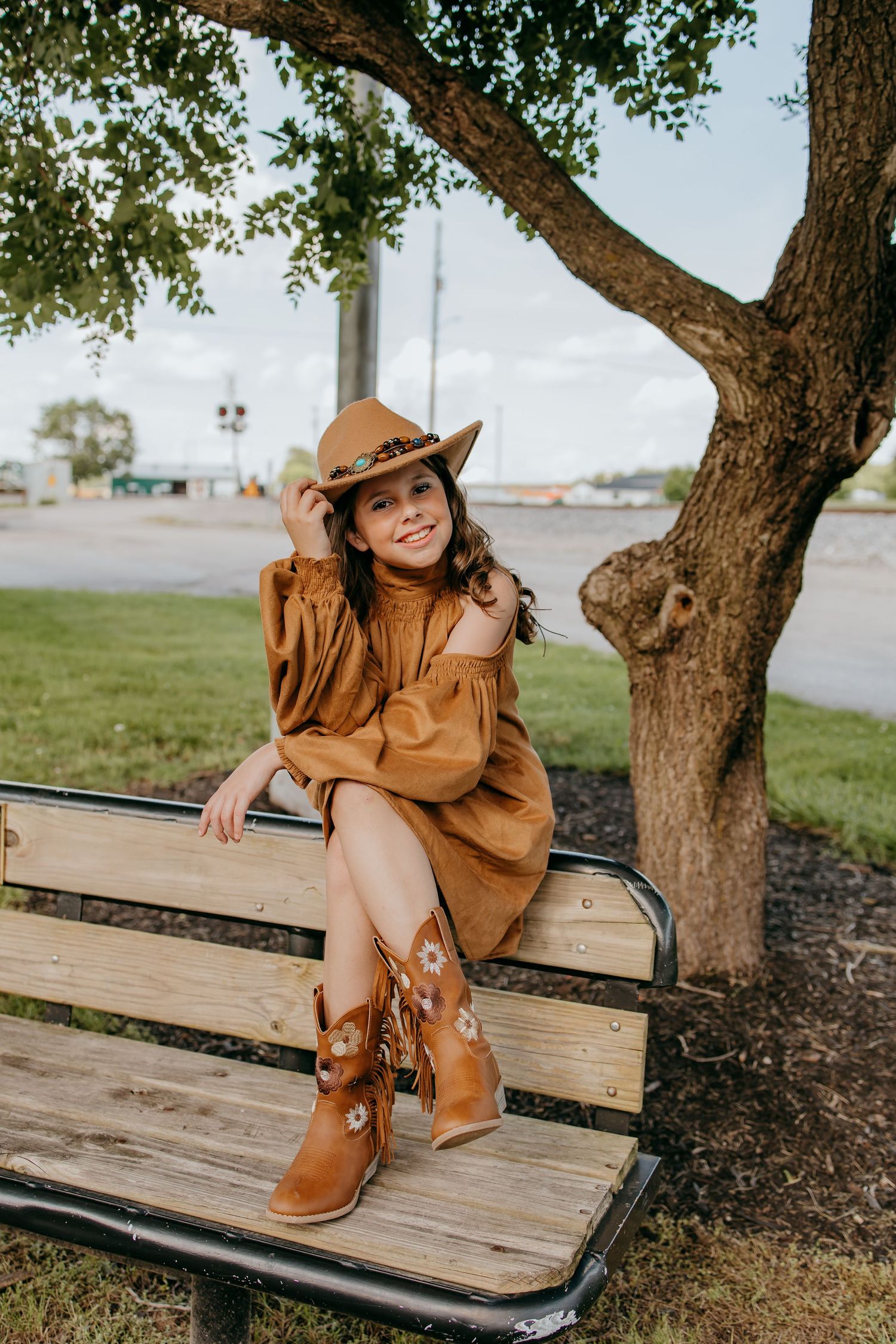 Young girl in brown cowboy hat, rust dress, and floral boots sits on a wooden bench under a tree.