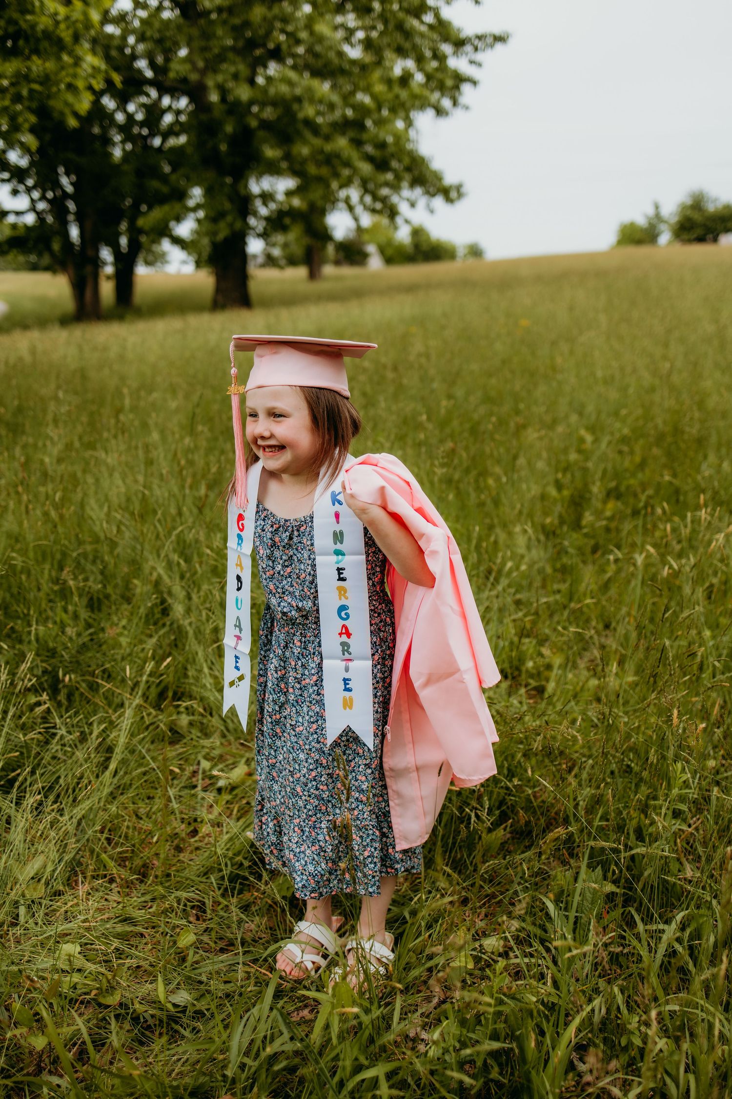 Young girl in pink kindergarten graduation cap and gown smiles proudly in a green field outdoors.