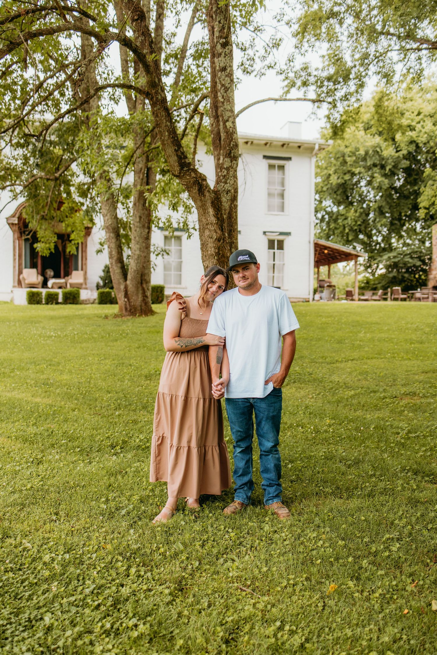 A couple stands hand-in-hand on green grass before a white farmhouse, woman in maxi dress, man in jeans and cap.