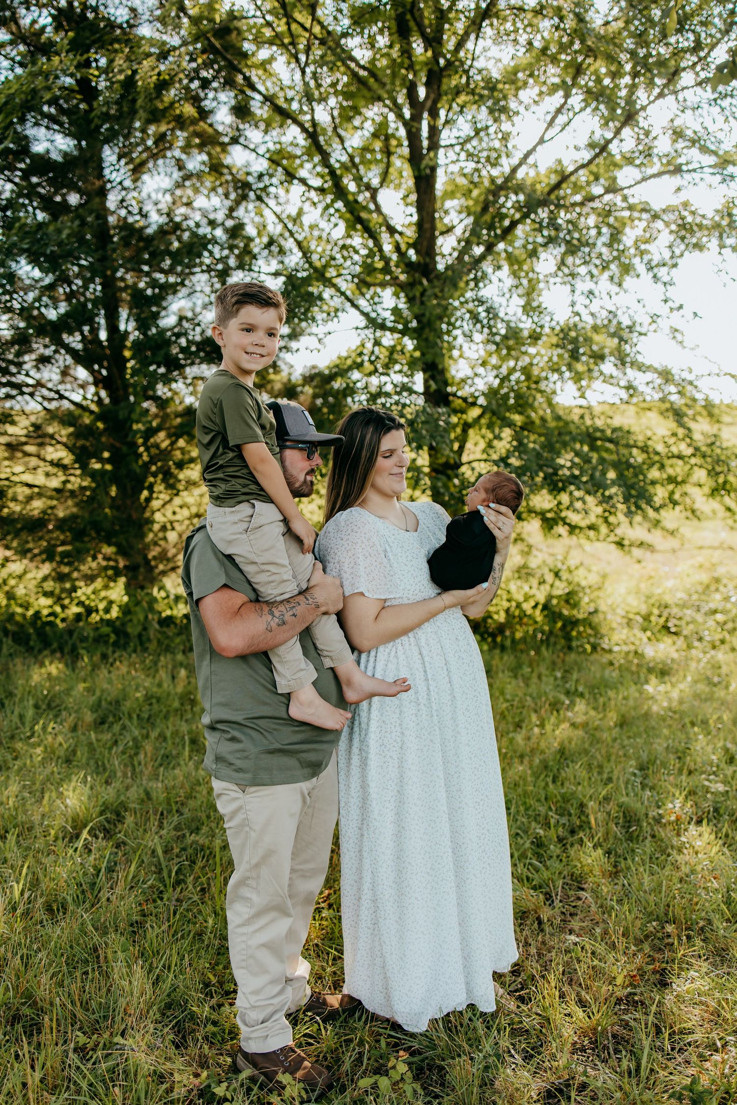 A family poses outdoors, dad holds son on shoulders while mom cradles newborn in white dress, surrounded by lush green trees.