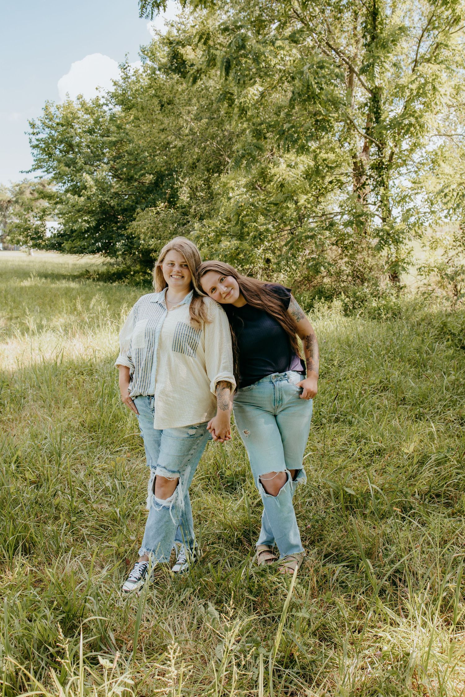 Two young women in ripped jeans pose together in a lush green field surrounded by trees on a sunny day.