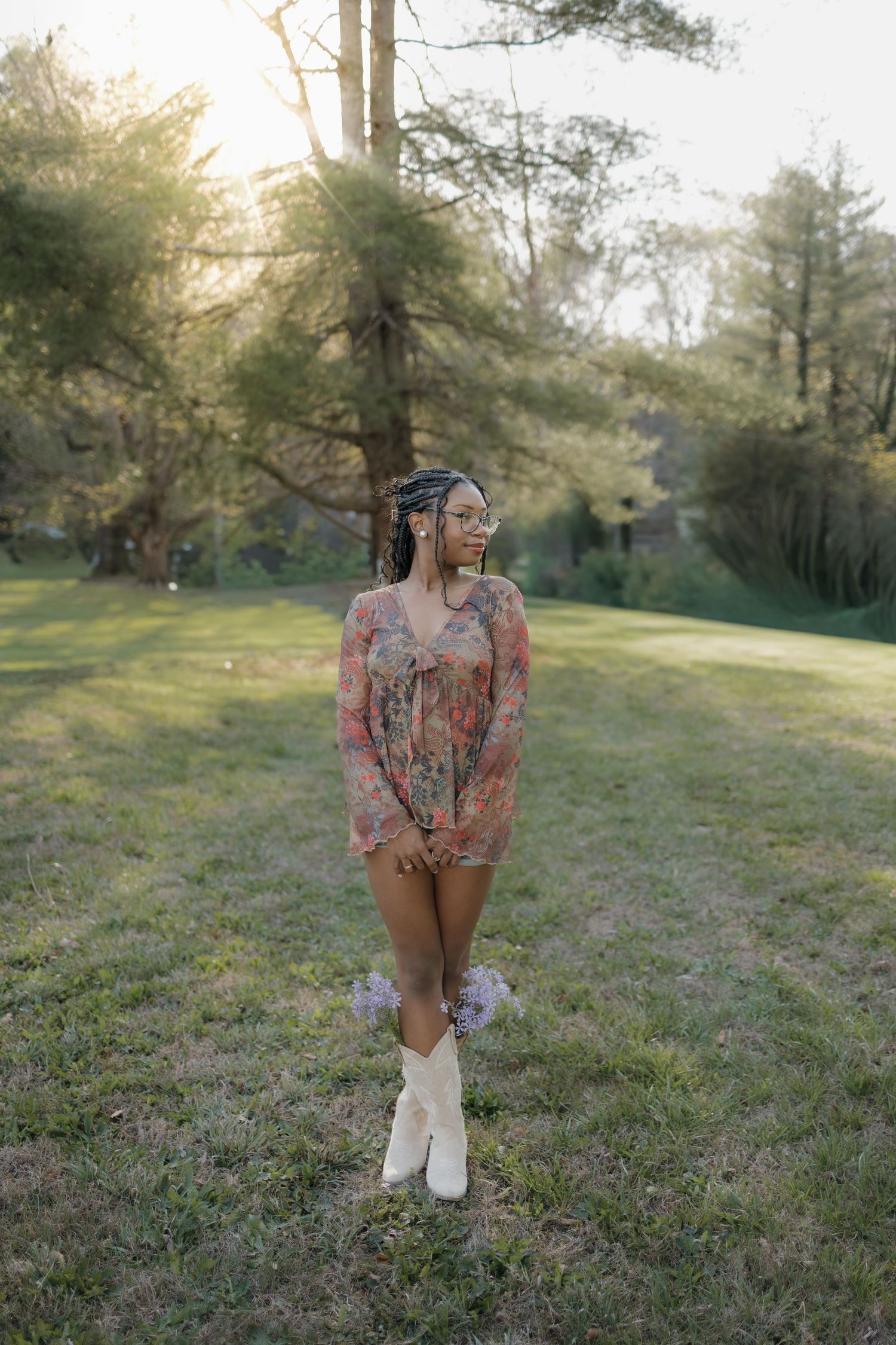 Young woman in floral romper and white boots poses outdoors in a sunlit park during golden hour.