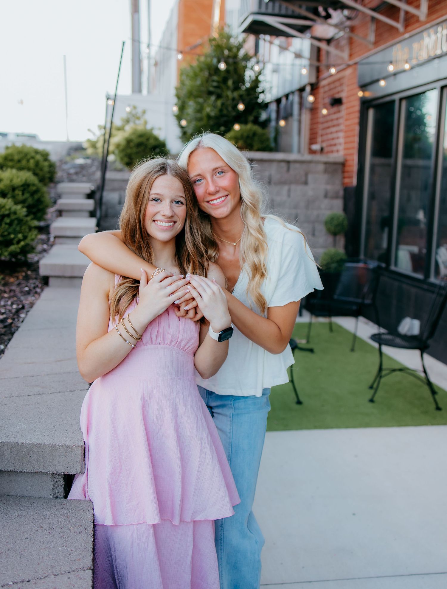 Two seniors  pose together outdoors, one in a pink dress, the other in a white top and jeans.