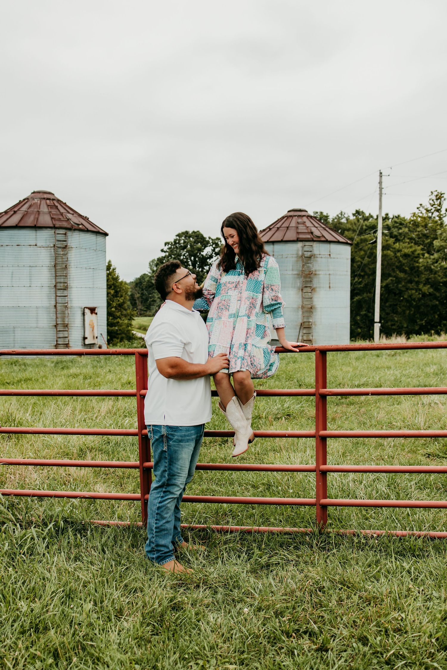 A couple shares a romantic moment by a red farm gate, with grain silos in the background on a lush green field.