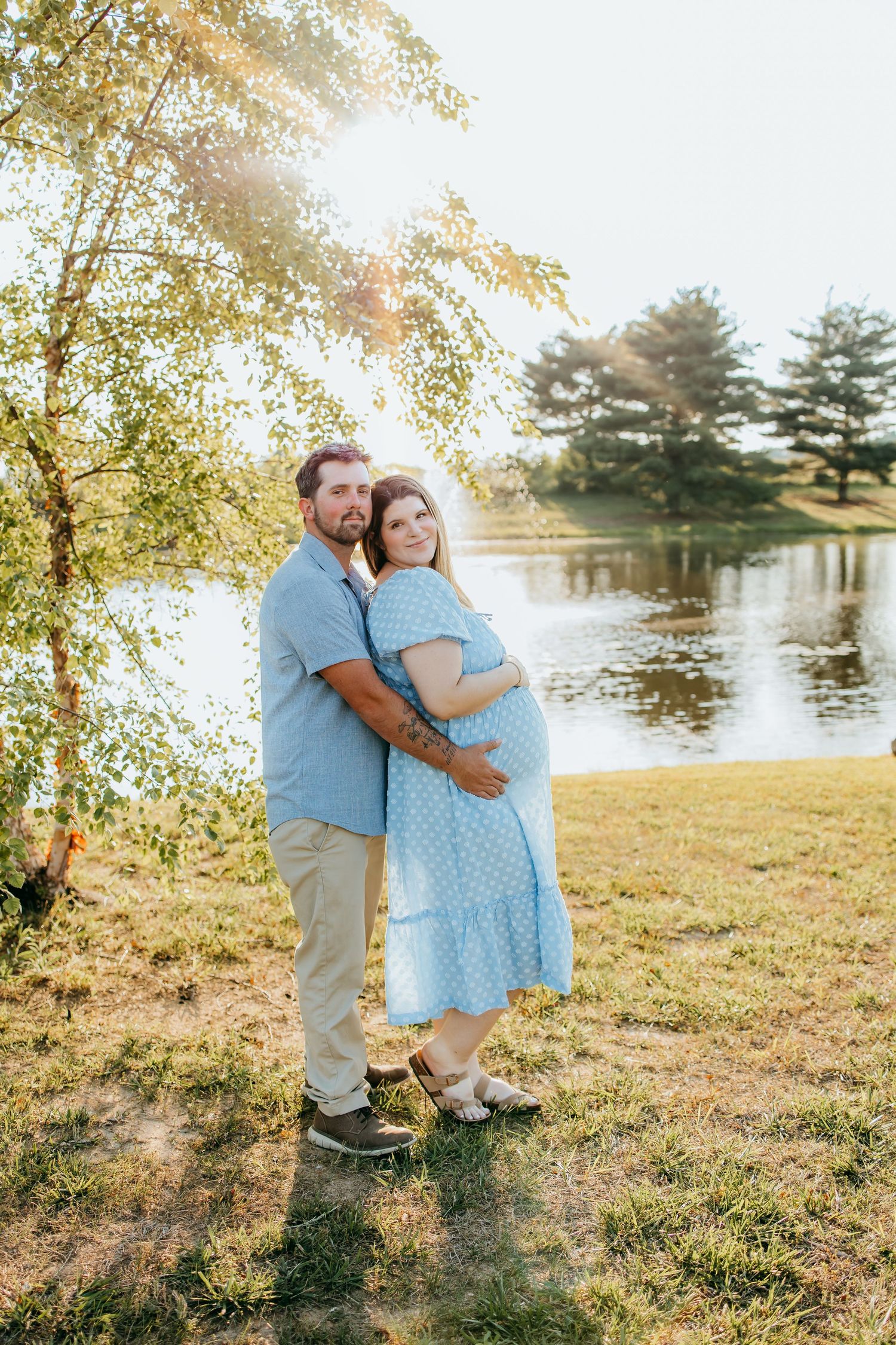 Pregnant couple embracing by a sunlit pond, woman in blue polka dot dress, man in blue shirt, golden hour outdoor maternity photo.