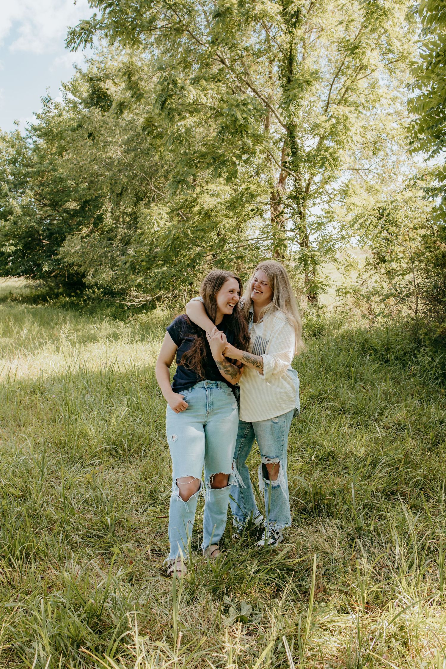 Two young women laughing and embracing in a sunny green field, wearing ripped jeans and casual tops.