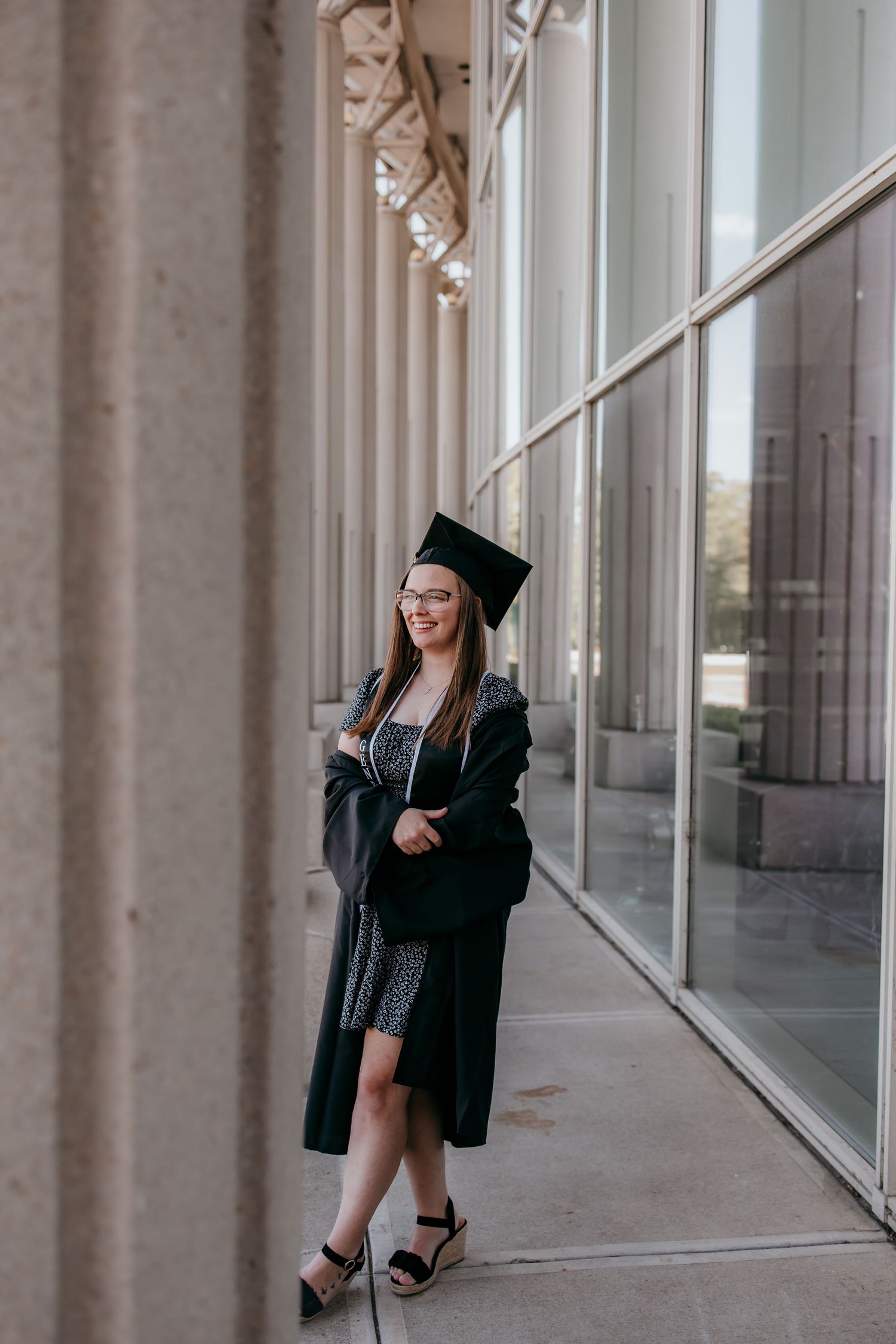 Young woman in black graduation cap and gown smiling outdoors near modern building columns.